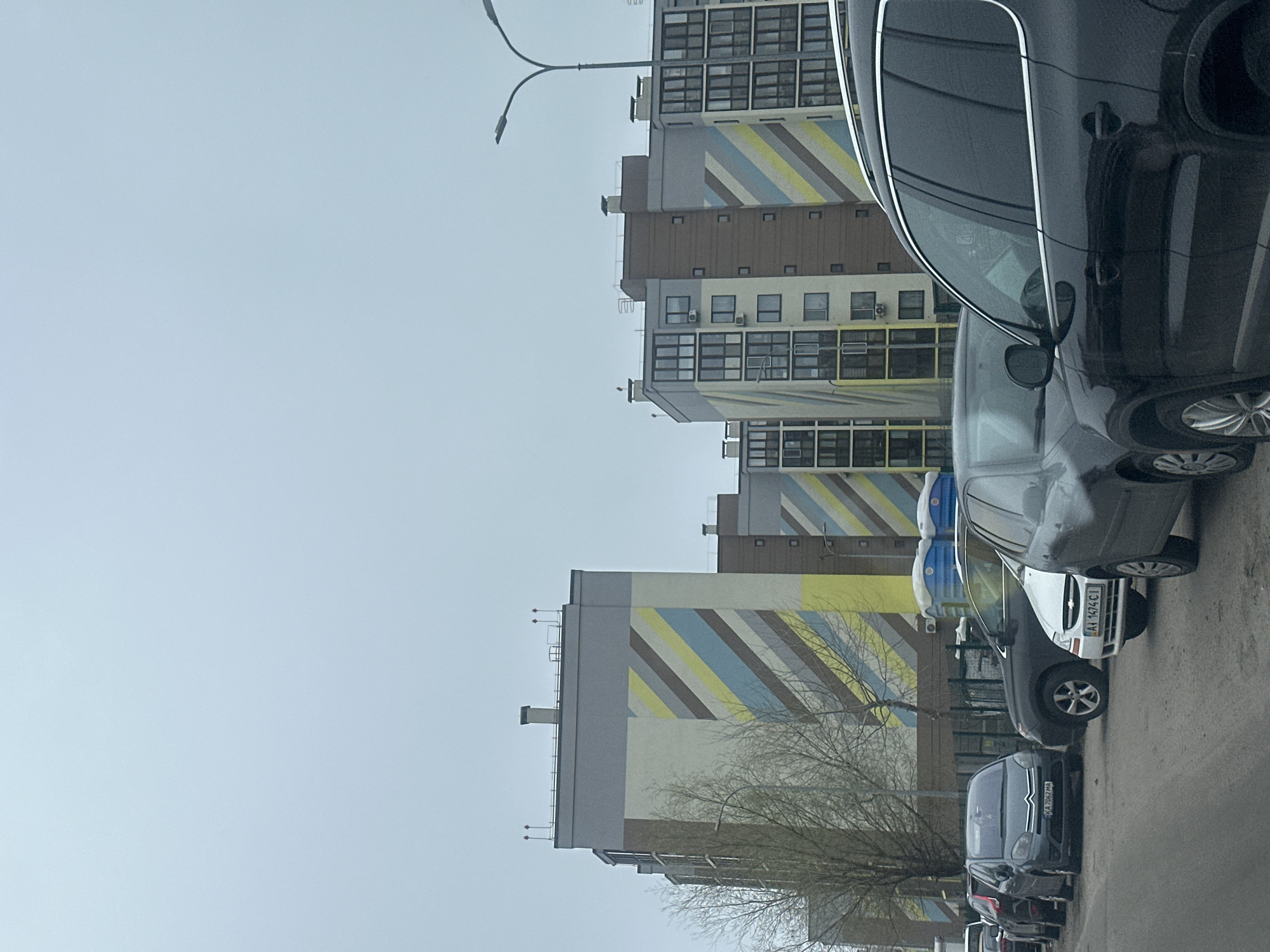 The image depicts a street scene in Kyiv, Ukraine. The focus is on a line of parked cars along a road next to a row of tall apartment buildings. The buildings are characterized by a modern design with geometric patterns, including angled stripes in blue, yellow, and beige. The sky is overcast. There are bare trees. The parked cars vary in model and color, including white, black, and grey. License plates are visible on some of the vehicles. A street light is seen in the background between the buildings.