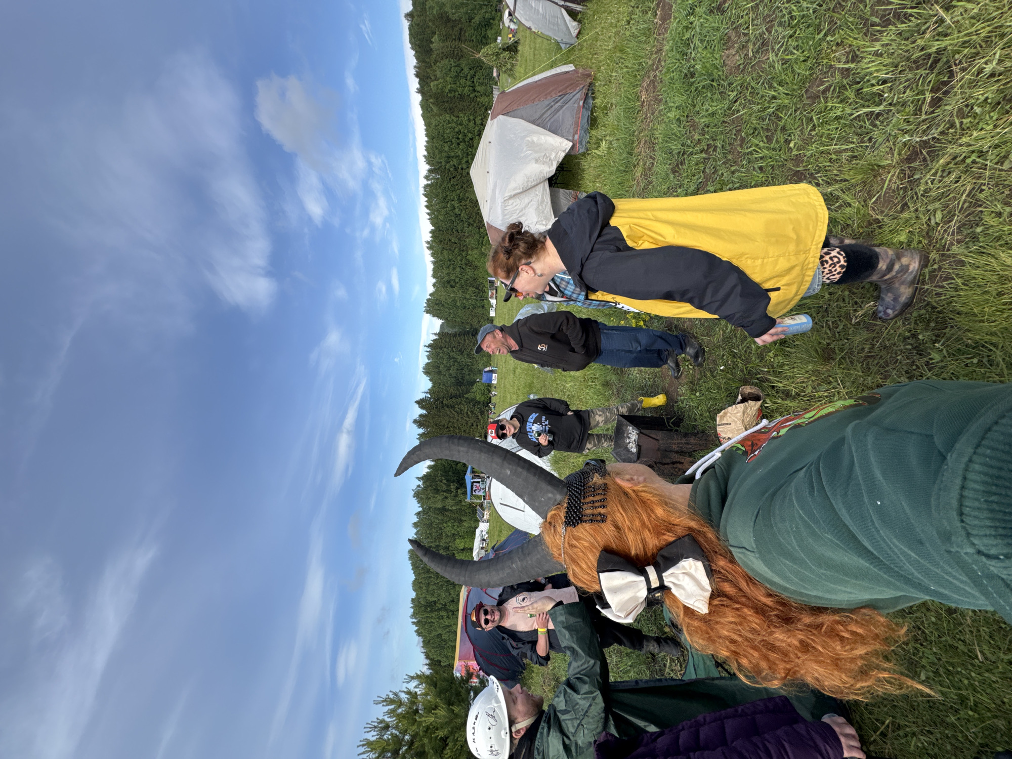 This image, presented as if correctly oriented, captures an outdoor gathering, likely a festival or camping event, in a grassy field in Bol'shaya Kivara, Russia. The scene is illuminated by bright daylight, under a vibrant blue sky dotted with scattered white clouds. A dense line of coniferous trees borders the field in the background, where several tents of various colors (grey, brown, white) are pitched.
In the immediate foreground, a person's head wearing a white hard hat is partially visible. Next to them, with their back mostly to the viewer, is a person with long, curly red hair adorned with large black horns and a prominent black and white bow. They are wearing a dark green long-sleeved top.
Moving from the left side of the mid-ground, a man in a blue cap, dark jacket (with a subtle white logo), and blue jeans appears to be smiling or laughing. To his right, a woman with reddish-brown hair tied in a bun, wearing sunglasses, a black top, and a bright yellow raincoat, holds a light blue can. She wears leopard print leg warmers peeking from her mud-splattered boots, suggesting the ground might be damp. Further right, another individual wearing a black hoodie, sunglasses, and camouflage pants stands near a wooden log or stump.
The event seems to involve casual socializing in a relaxed outdoor environment. People are dressed for comfort and potentially variable weather, with raincoats and boots visible. A paper bag on the ground near the wooden log displays the partial text "SKA". The overall mood appears genial.