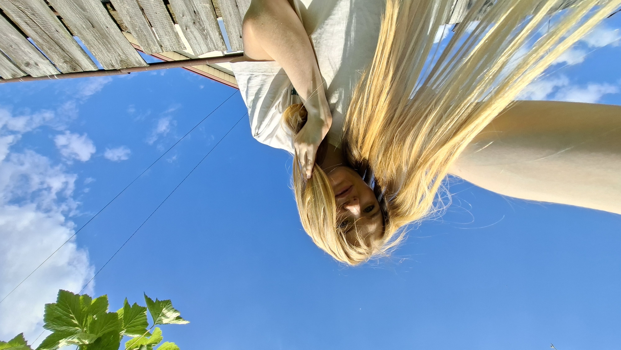 A young woman with long blonde hair is captured in a low-angle, outdoor selfie. She is wearing a light-colored t-shirt and her left arm is extended towards the camera, obscuring part of the frame. Her right hand is resting on her chin, and her gaze is directed slightly away from the camera. The background is dominated by a bright blue sky with scattered white clouds. A wooden fence runs along the bottom right of the image, and the roofline of a building with a light brown facade is visible behind the fence. Green leaves from a nearby plant hang from the top left corner of the image. Power lines are visible in the upper portion of the sky. The scene appears to be set in a rural or suburban area, possibly within the city of Bolshoy Bukor, Russia, given the context.