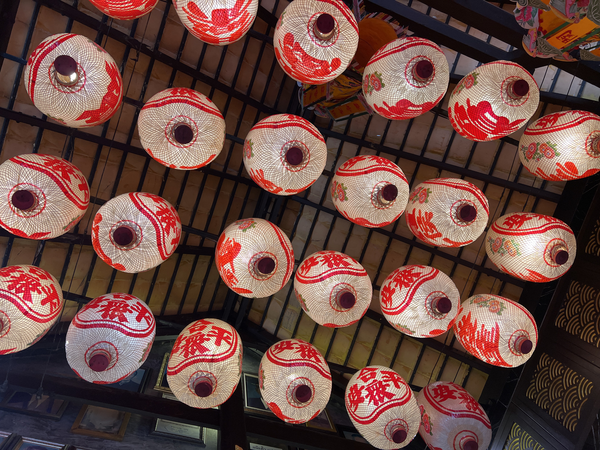 A low-angle, close-up shot captures a collection of traditional lanterns hanging from a ceiling in Baan Hua Thanon, Thailand. The lanterns are predominantly white with intricate red Chinese characters and floral designs adorning their surfaces. The woven texture of the lanterns is visible, and some emit a warm, soft light from within. They are suspended at varying heights, creating a visually dynamic arrangement against the backdrop of a dark, tiled ceiling and wooden beams. To the left, portions of framed pictures are discernible on a wall. The overall impression is one of festive decoration and cultural ambiance.