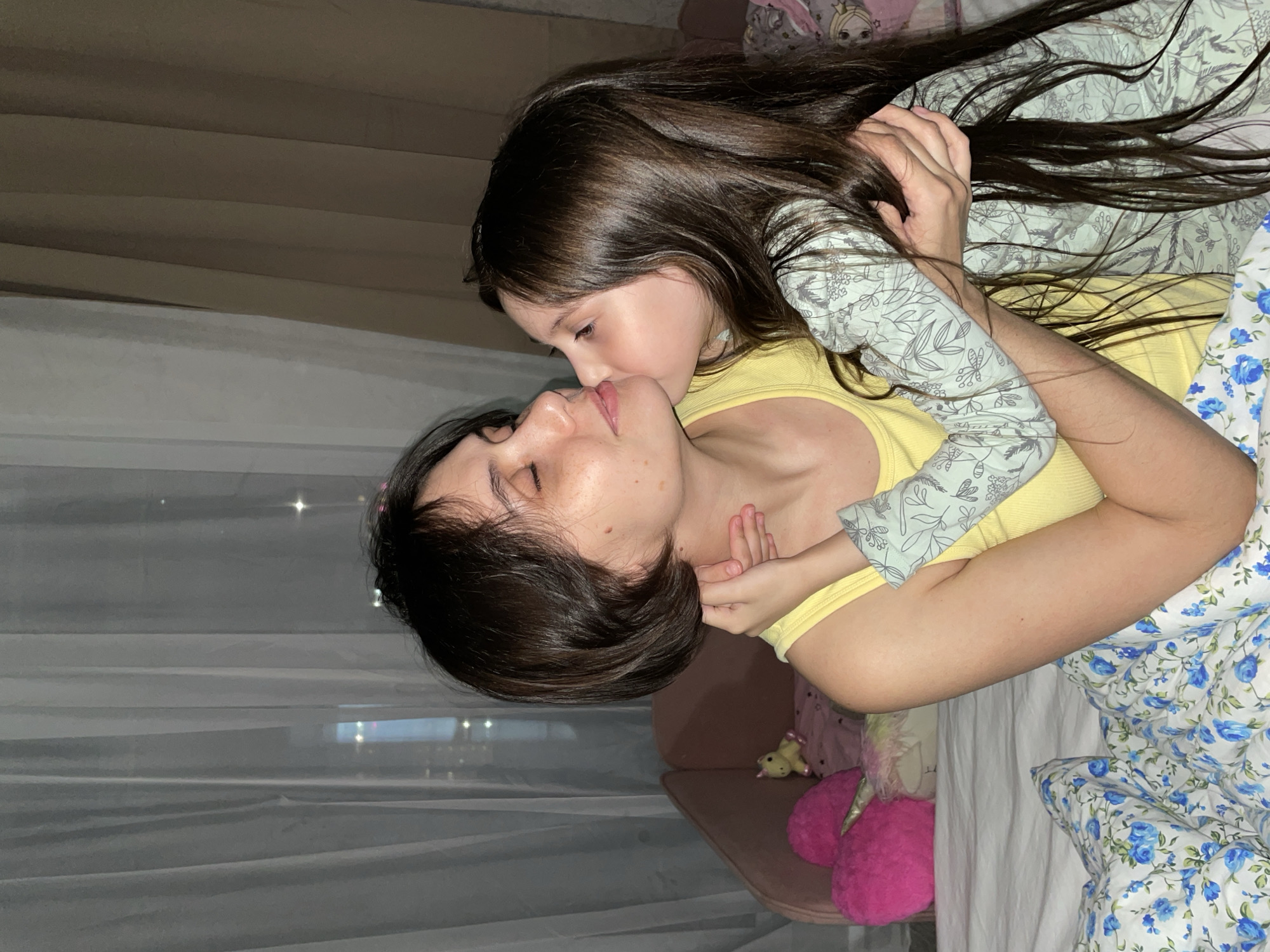 A woman and a young girl are lying on a bed in a room. The woman is lying on her back, wearing a yellow tank top. The girl is lying on top of the woman, facing her, and appears to be kissing the woman's lips. The girl has long dark hair and is wearing a light-colored, long-sleeved pajama top with a floral pattern. The bed has a white sheet and a blue and white floral patterned blanket. In the background, there is a window with sheer white curtains and a glimpse of a city view at night. The location cannot be determined to be Izhevsk, Russia from the visual information in the image.