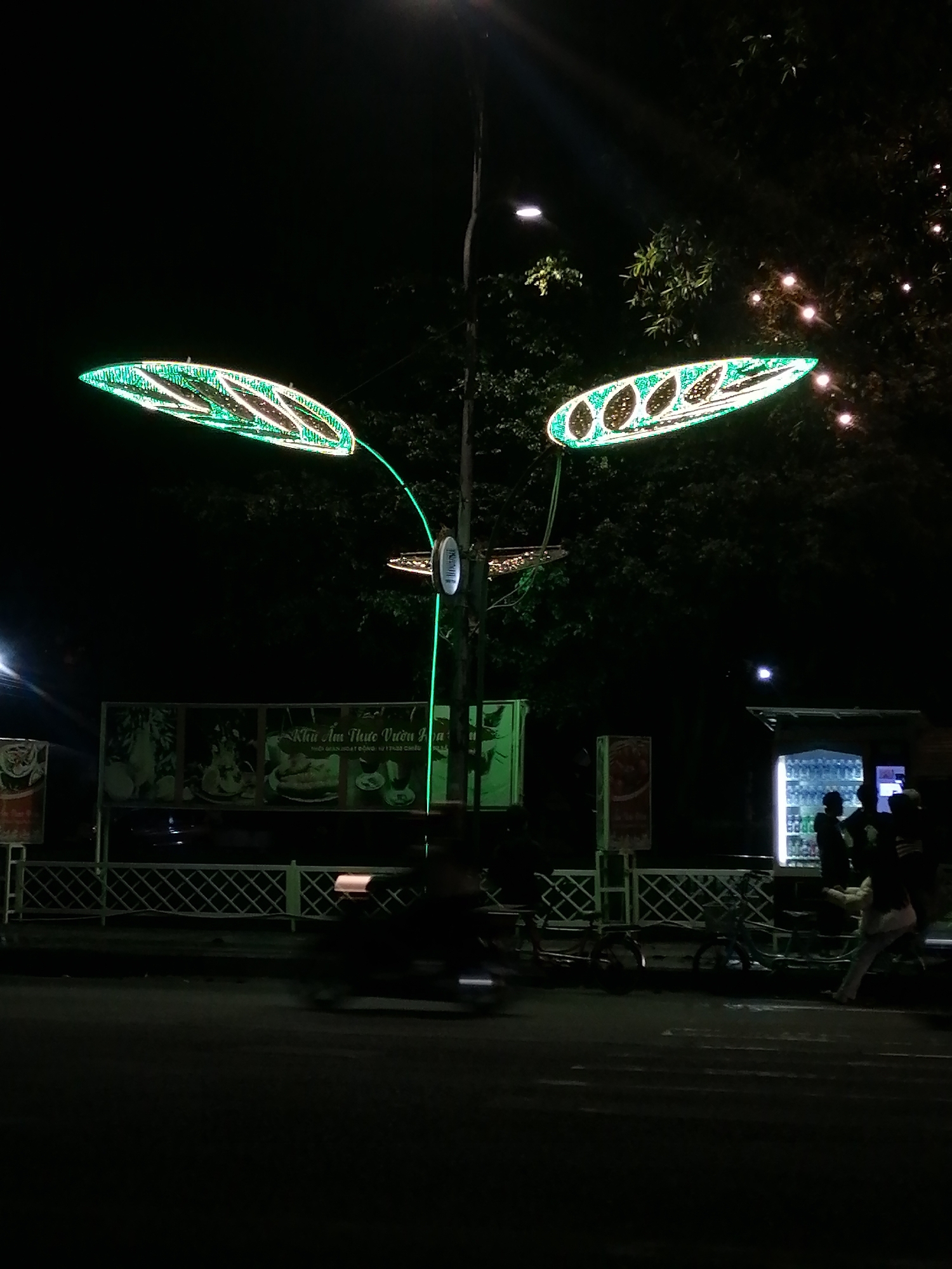 The photograph, taken at night, shows a street scene likely in Da Lat, Vietnam, given the presence of Vietnamese signage. A street lamp with three leaf-shaped green neon lights illuminates the area. Below the lamp, there is a sign in Vietnamese, potentially advertising a restaurant ("Khu Am Thuc Vuon"). A low white fence runs along the edge of the sidewalk. Several blurred figures are visible, including a person on a motorbike passing by, and a group of individuals standing near a brightly lit beverage vending machine. The background features trees with some small white lights strung through the branches.