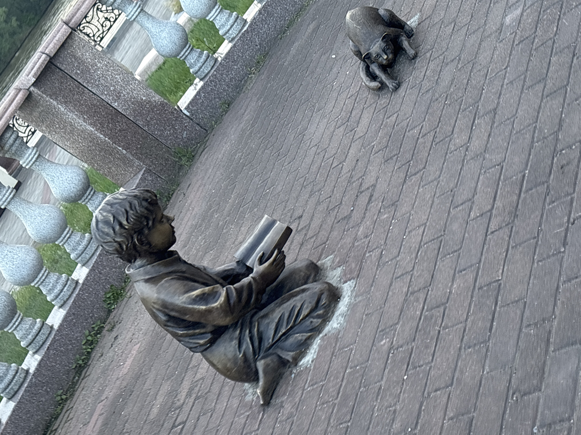 The image shows a bronze sculpture of a child kneeling and reading a book, positioned on a cobblestone pathway. To the right of the child, a bronze sculpture of a cat is depicted in a crouched, playful stance, facing the child. In the background, there is a stone balustrade with decorative elements, green grass, and a body of water. The scene appears to be in an outdoor public space, possibly a park or garden, within an unknown city.