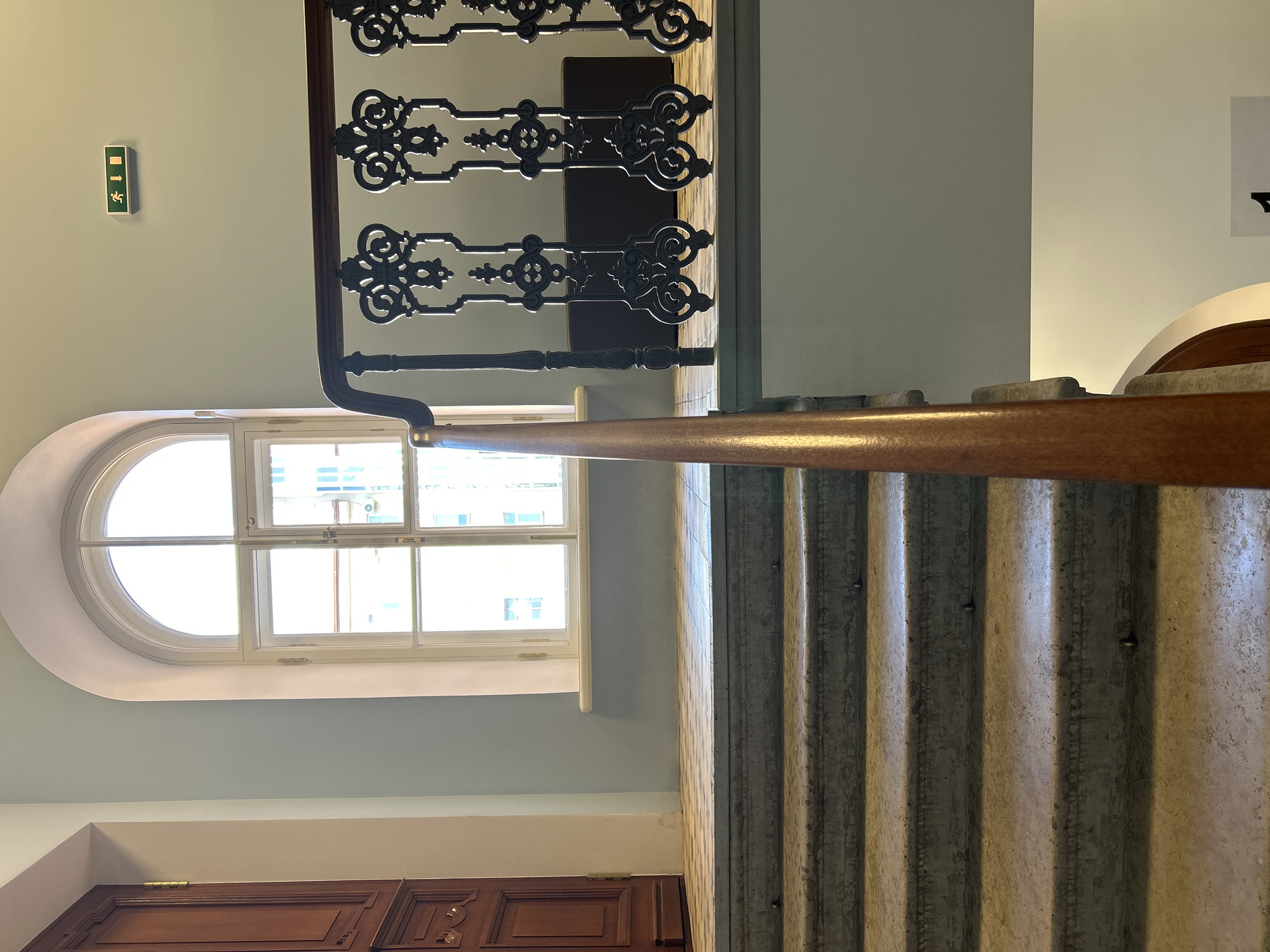 The image displays an interior view of a staircase, oriented vertically due to a 90-degree clockwise rotation. On the right side, a smooth, polished wooden handrail runs vertically, guiding the ascent or descent of the staircase. The stairs themselves, visible on the right, are made of light-colored stone or concrete, featuring distinct horizontal lines and visible texture from wear.
To the left of the handrail, a striking, ornate black wrought iron railing stands vertically. This railing is richly decorated with elaborate scrollwork and stylized floral patterns, adding a sense of grandeur to the space. Further to the left, a large, arch-topped window with white frames and multiple panes allows abundant natural light to enter, indicating a daytime scene. Through the window, faint outlines of buildings can be discerned.
Above the window, on a light-colored wall, a green rectangular emergency exit sign is mounted. It prominently features a white pictogram of a running person and an arrow pointing upwards, indicating the exit route in this orientation.
At the bottom of the image, beneath the window, a section of dark wooden paneling or a decorative door is partially visible, showing some carved details. The walls are painted in a soft, neutral tone, possibly a pale green or off-white. No individuals are present in the photograph. The scene suggests an older, architecturally significant building, likely public or institutional, characterized by its classic design and functional safety elements.