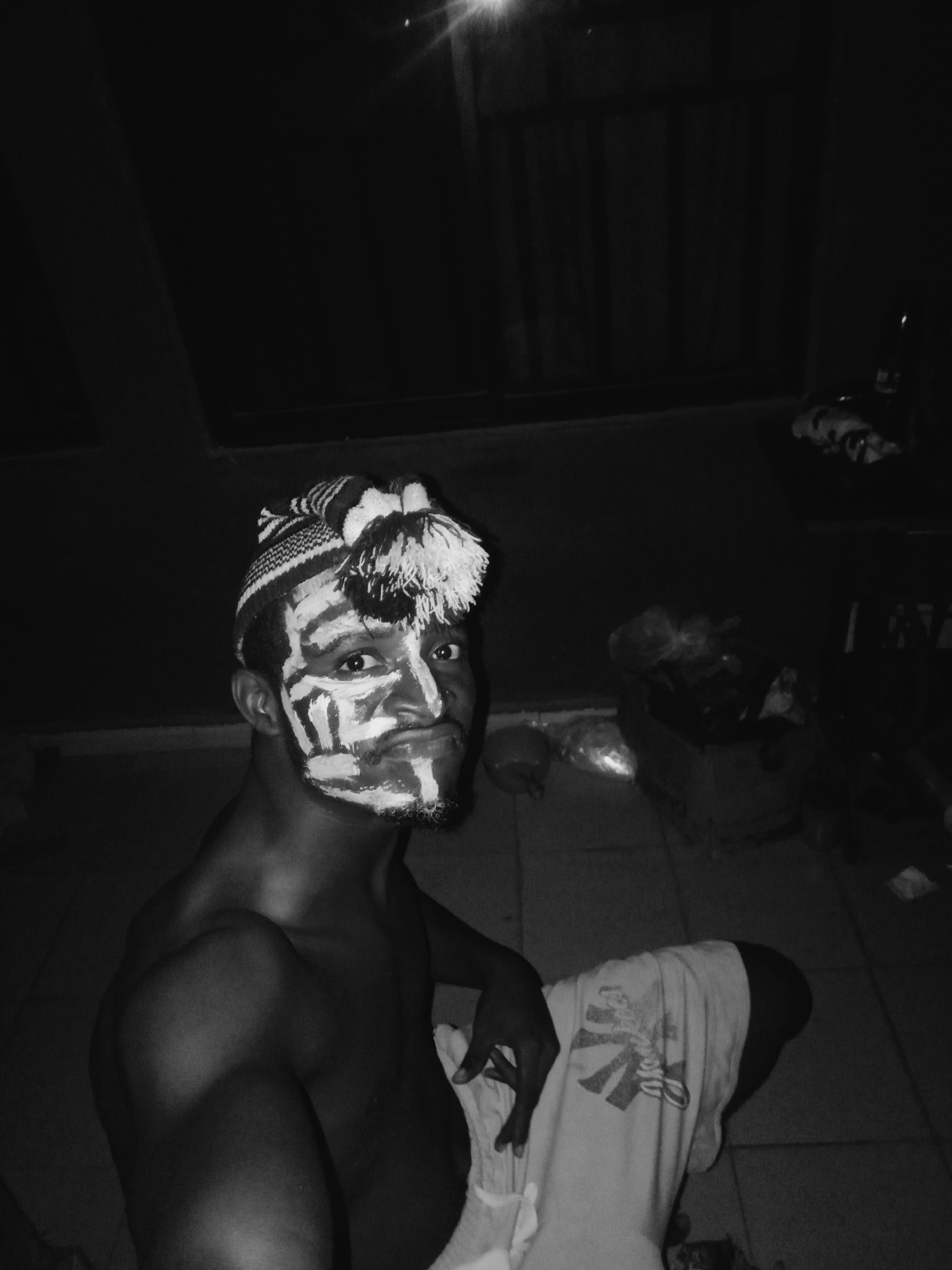 A young man, likely of African descent, is the central subject in this black and white, low-light photograph. He is bare-chested, revealing his shoulders and upper torso. His face is strikingly adorned with bold, white paint applied in abstract, geometric patterns across his forehead, cheeks, and nose. He has a short beard. On his head, he wears a striped, knitted beanie hat with a prominent pompom on the side. A light-colored garment, possibly a wrapper or shorts, is visible around his lower body, featuring an indistinct design.
The man looks directly at the camera with a direct gaze and a slight smirk or playful pout. His body is turned slightly, with his right shoulder prominent. The setting appears to be a dimly lit indoor or covered outdoor space, likely at night, indicated by the strong artificial light source in the upper left that creates a lens flare. The floor is tiled. In the background, indistinct objects such as a cardboard box and several plastic bags or wrapped items are faintly visible on the right, suggesting a utilitarian or storage area.
The facial painting, coupled with the location context of Onitsha, Nigeria, suggests a cultural event, performance, or ceremonial preparation. The monochromatic palette and stark lighting create a high-contrast and somewhat mysterious mood. No discernible text is visible in the image.
