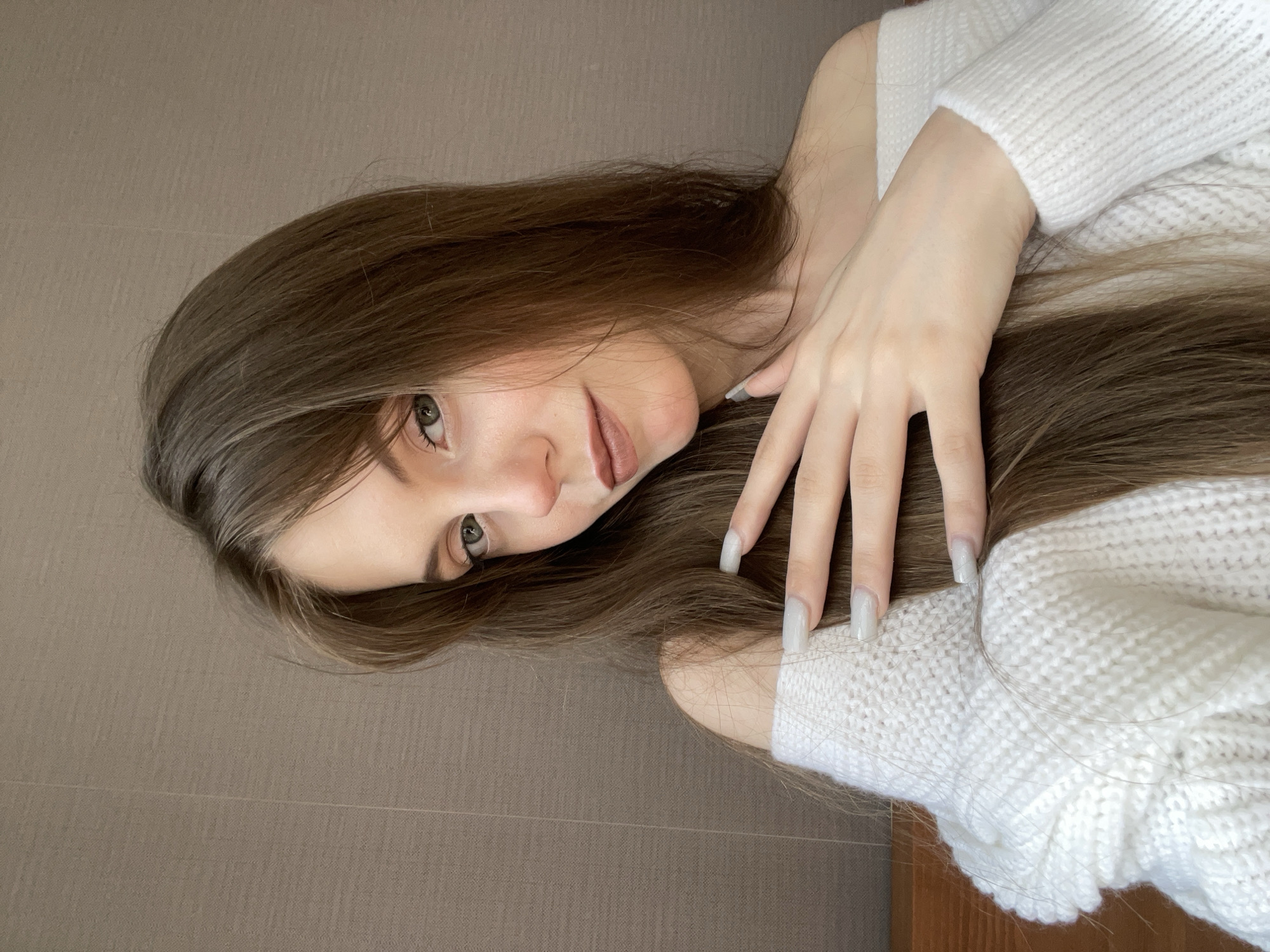 A young woman with long, light brown hair is positioned in front of a light brown wall, possibly indoors. She is wearing a white sweater and her hand is gently resting on the fabric. Her face is tilted towards the viewer, her eyes are light colored. The scene appears to be indoors. There is no definitive evidence to place this photo specifically in the city of Ufa, Russia.