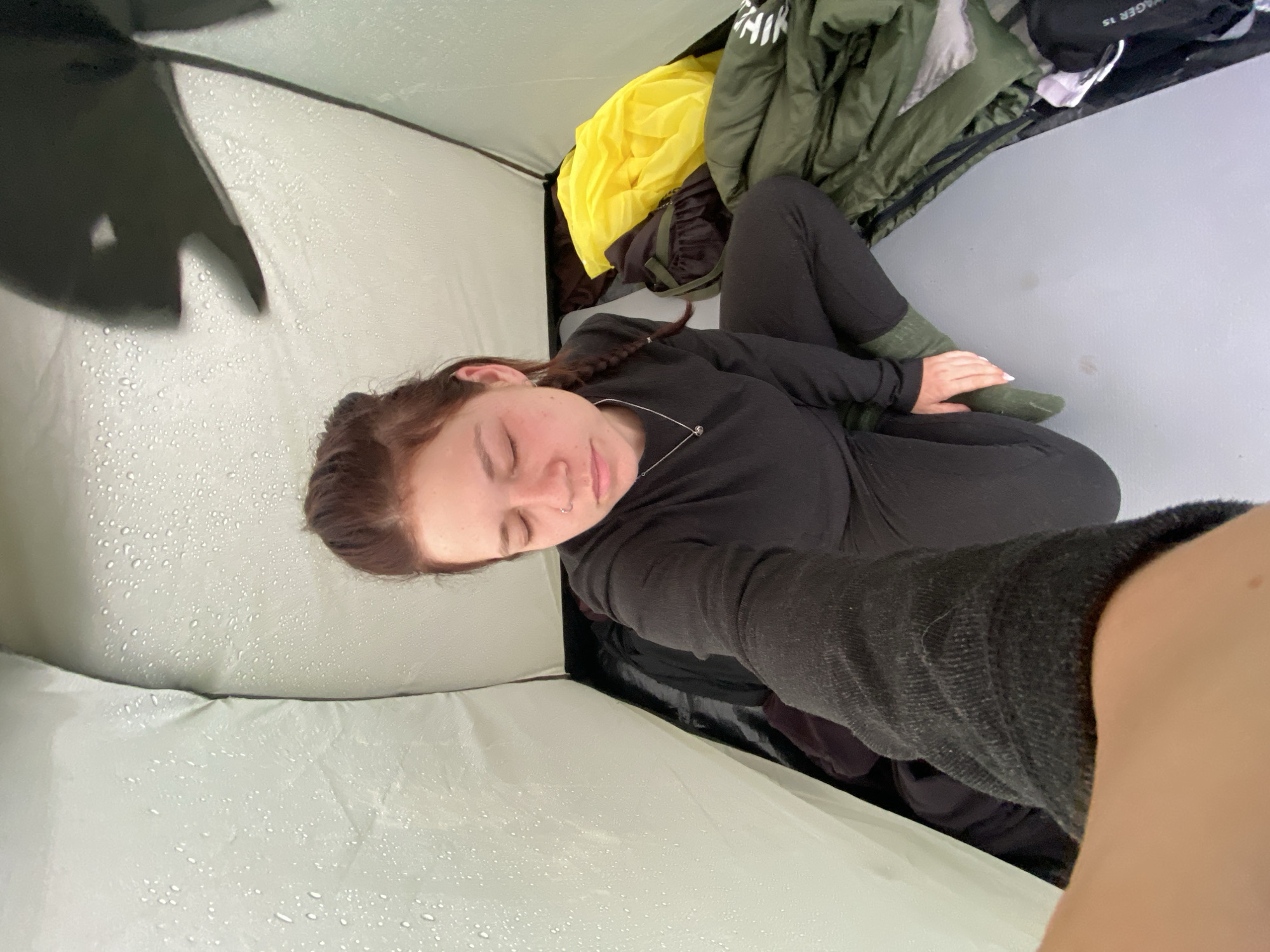 A high-angle, medium shot shows a young woman sitting inside a tent, with her eyes closed and her head tilted down. She has auburn hair pulled back in a braid and is wearing a black long-sleeved shirt and black pants, with green socks. The tent's interior is visible, with water droplets on the fabric. To her right are camping supplies, including a green sleeping bag and a bright yellow bag. The floor of the tent is a light grey material. The scene is set in the natural landscape of Абзаново, Russia, which is evident from the context of camping.