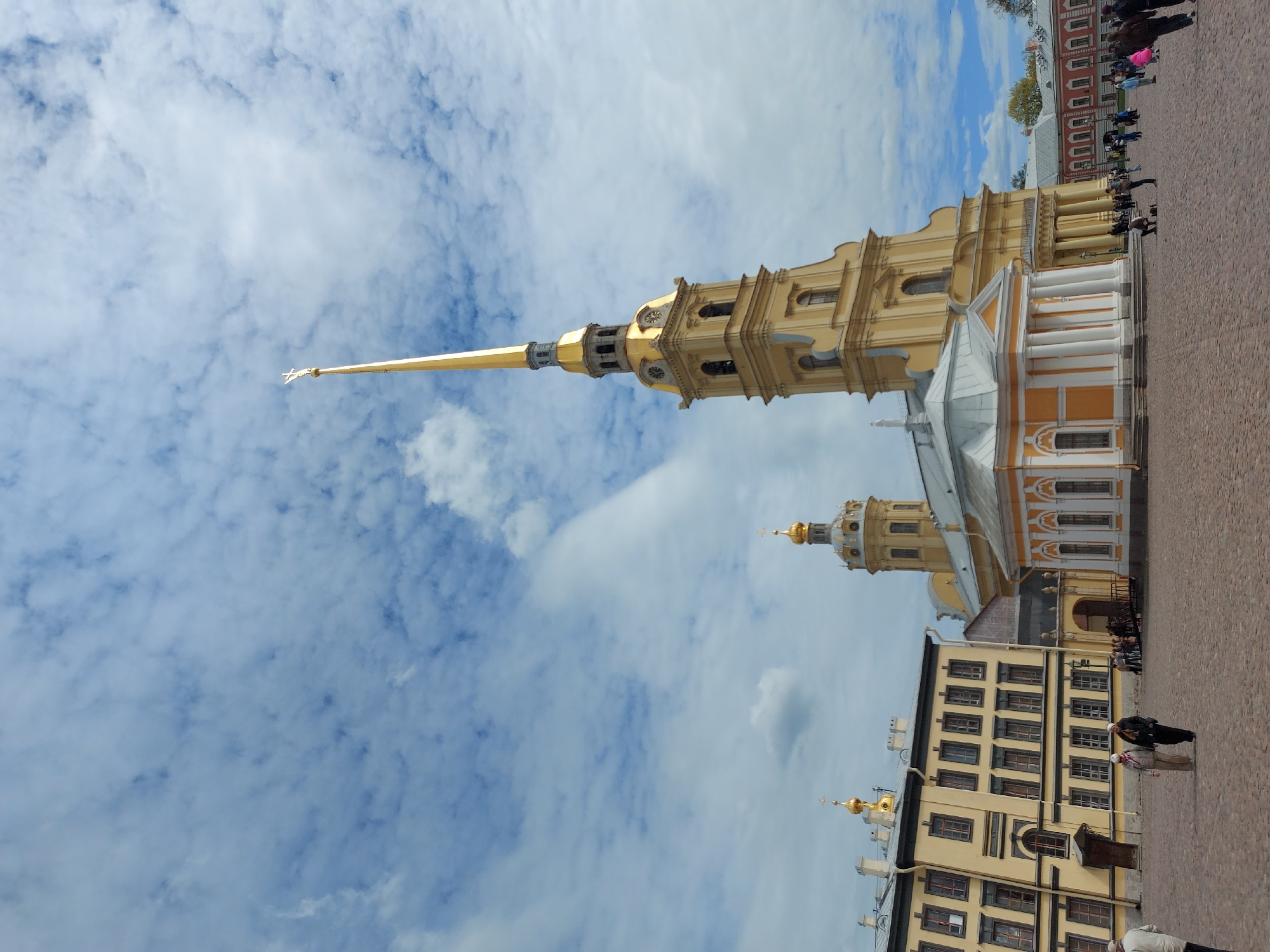 The image shows a view of the Peter and Paul Cathedral in Saint Petersburg, Russia. The cathedral's tall, golden spire dominates the frame against a backdrop of a sky with white clouds. The building is multi-tiered, with a pale yellow facade, and adorned with white columns. Other buildings are seen as well. People are visible on the brick-laid ground.