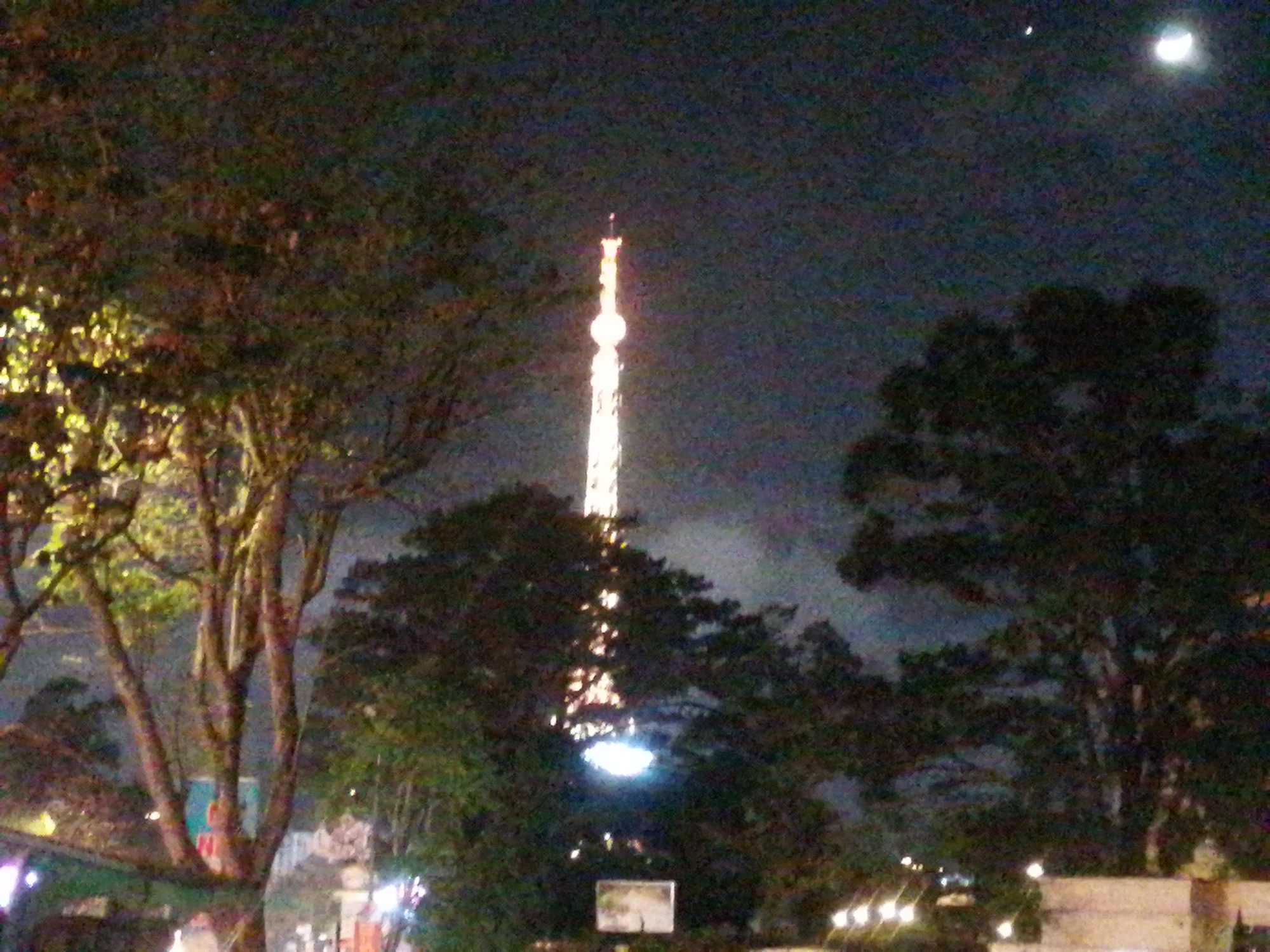 At night in Da Lat, Vietnam, a brightly illuminated, tall tower rises through trees. A crescent moon and a faint celestial object are visible in the dark sky above. Trees are in the foreground on both sides. The illumination indicates it is likely the Da Lat television tower. Streetlights are visible at ground level. The image is somewhat grainy, suggesting a low-light photography condition.