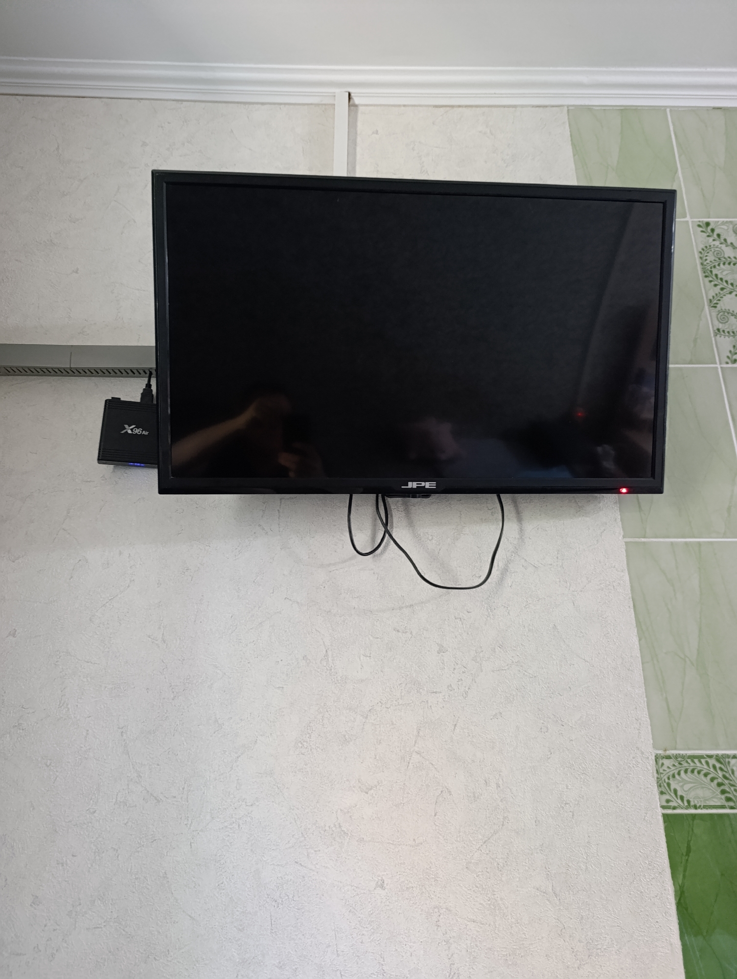 The image shows a flat-screen television mounted on a wall. The TV has a black frame and the screen is dark, reflecting a person's hand. The logo "JPE" is visible on the bottom bezel of the TV. To the left of the TV, a black media box with the logo "X96 Air" is attached to the wall. Cables are connected to the media box and the TV. The wall behind the TV has a textured, light-colored finish, and to the right, there are green tiles with a decorative pattern. The scene is indoors, likely in a residential setting. There is no information in the image to indicate the location in Roghi, Moldova.