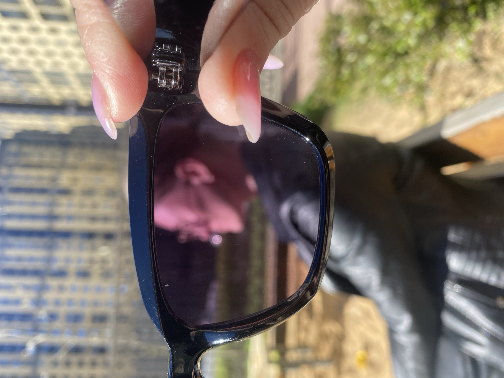A hand with long, manicured nails holds up a pair of black sunglasses, partially obscuring the view through the lenses. A reflection can be seen in the lens. In the background, there appears to be a blurred outdoor scene with elements suggesting a patio setting, and greenery. The scene is taken outdoors, with good lighting. The focus is sharp on the sunglasses and the hand. It's not possible to determine the exact location or any specific actions from the provided image.