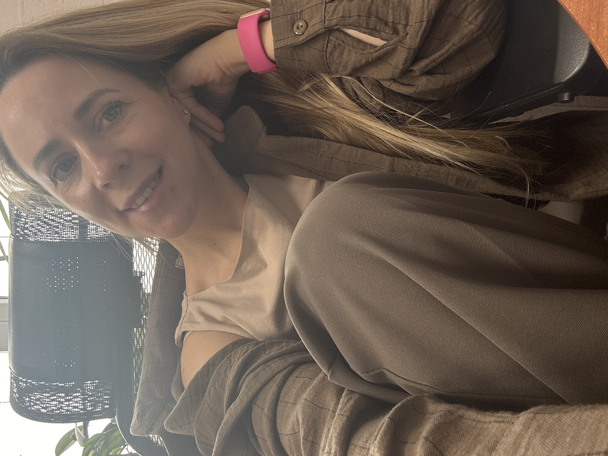 A woman with blonde hair, wearing a pink wrist device, a brown collared shirt, and a beige top, is sitting in a chair. She is smiling slightly and resting her head on her hand. The background includes a potted plant with a decorative black metal container. Based on the known location, this image was likely taken indoors in the city of Chaikovsky, Russia.