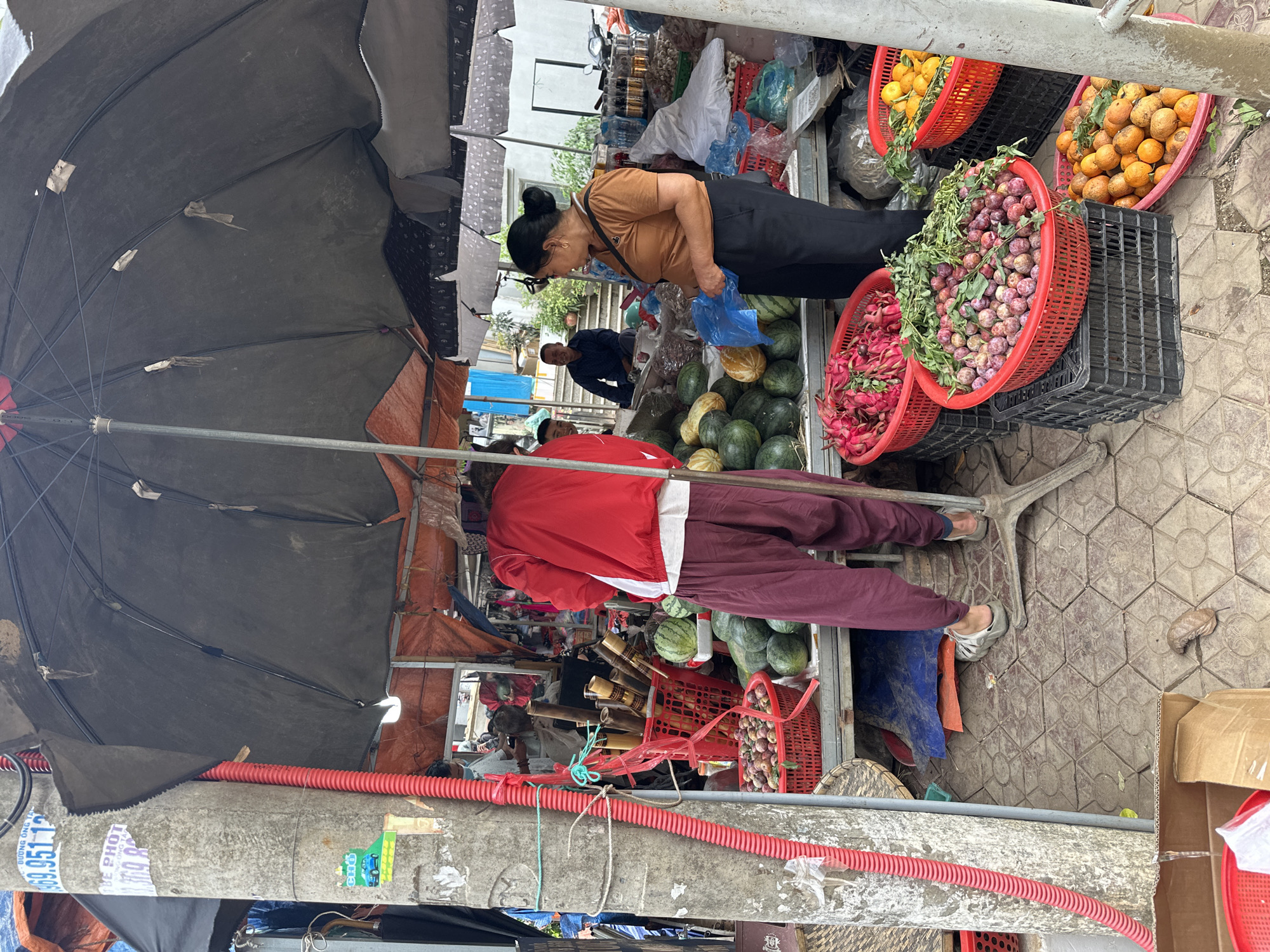 A street market scene unfolds in Bắc Yên, Vietnam, under the shade of large umbrellas. A person in a red jacket and maroon pants stands facing away from the camera, interacting with a vendor. The vendor, a woman in a brown shirt and black pants, stands behind a display of fruits, holding a blue plastic bag. In front of them, a variety of produce is arranged in red baskets and on a table, including watermelons, what appear to be plums, and oranges. Further in the background, other market stalls and people can be seen. The ground is paved with hexagonal tiles. A utility pole with red and green tubing attached stands to the left of the frame, displaying various stickers and notices.