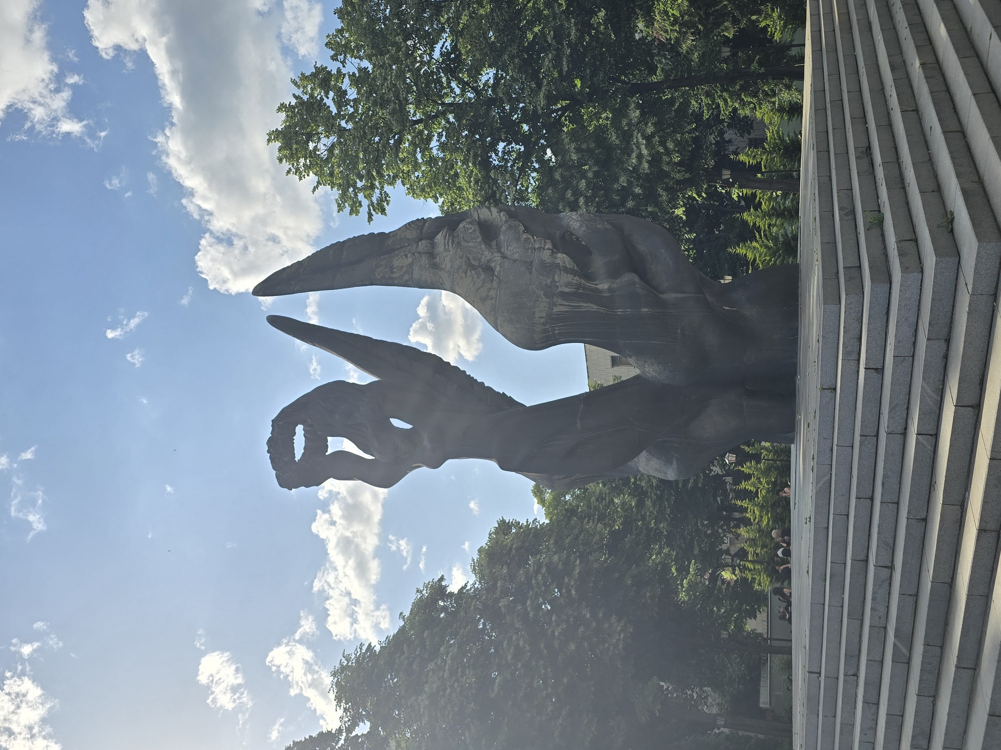 Here is a forensic description of the image:
The image depicts a sculpture in an outdoor setting. The sculpture features an abstract form that appears to have human figures. The sculpture is made of a dark material. It is set against a backdrop of a blue sky, clouds, and the tops of tall, green trees. There is a set of stairs to the right of the sculpture. The location is in a city of Unknown, Unknown.