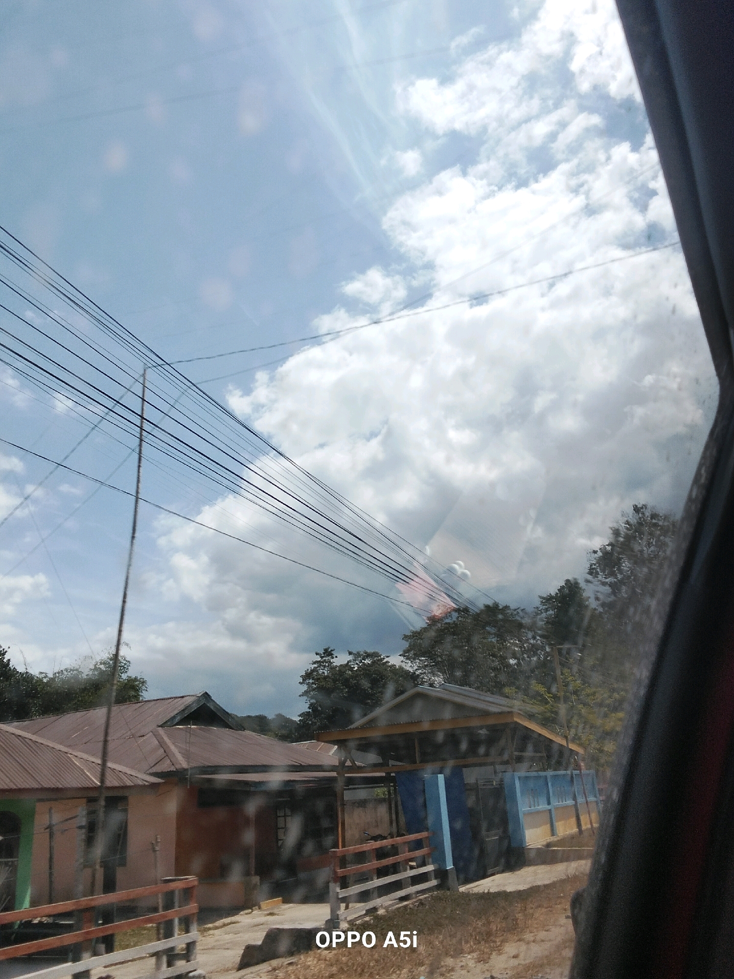 The image captures a street view in Andadowi, Indonesia, under a partially cloudy sky. The foreground is dominated by a series of dark power lines stretching diagonally across the frame. To the left, two houses with corrugated metal roofs are visible. One house is painted a pale pink and green, while the other is a reddish-brown. A wooden fence with red and white railings runs in front of these houses. Further to the right, another structure with a light-colored roof and blue and yellow walls is partially visible, with trees and foliage behind it. The overall impression is of a residential area with basic infrastructure. The bottom right corner of the image contains a black watermark, "OPPO A5i."