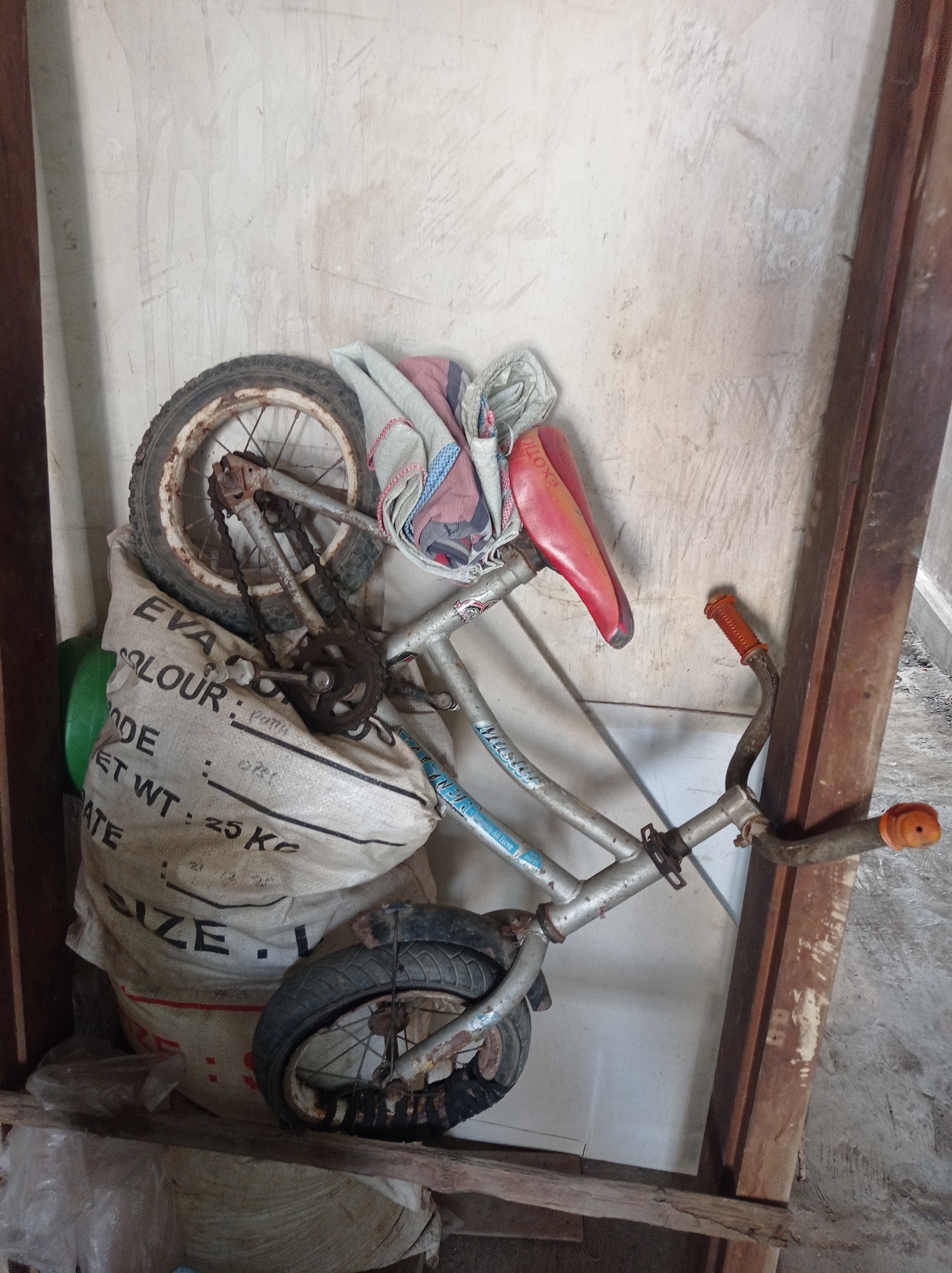 A close-up, eye-level shot depicts a worn, red bicycle seat resting on a silver bicycle frame, partially obscured by a large, off-white woven bag. The bag, marked with black text that includes "COLOUR" and "NET WT: 25 Kg", appears to be filled with something, causing it to bulge. A bicycle wheel with a rusted rim and spokes is visible at the top of the bag, and another wheel with a deflated tire is positioned at the bottom. The bicycle frame shows signs of rust and wear. Orange grips are present on the handlebars. The background is a textured, pale wall, possibly made of concrete, with a vertical wooden beam on the right side of the frame. The scene appears to be an interior space, possibly a storage area, with a floor of concrete or similar material visible at the bottom. The lighting is soft, suggesting an indoor environment with ambient light.