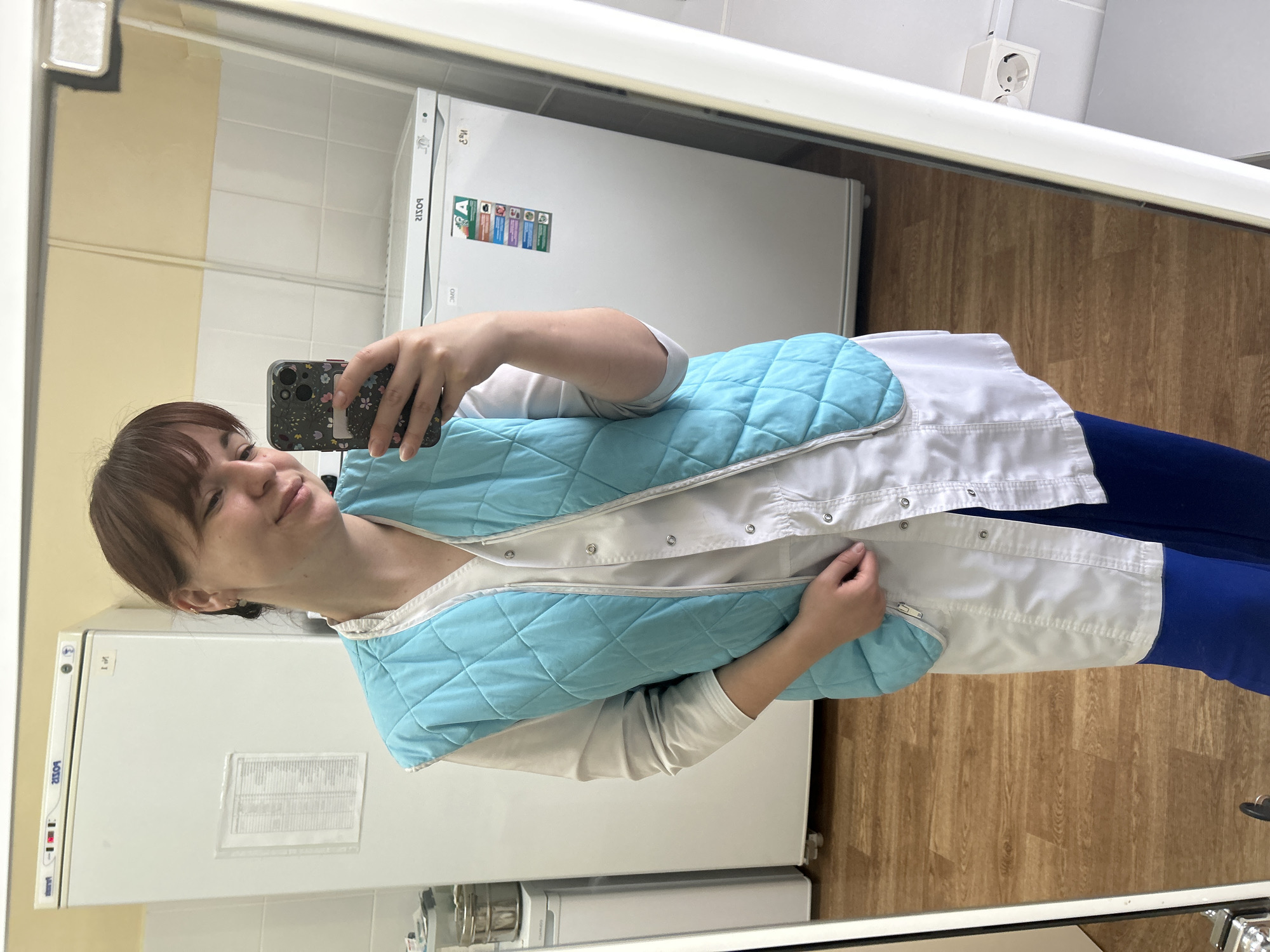 A young woman with light brown hair and bangs takes a mirror selfie in an indoor setting. She is wearing a white long-sleeved shirt, a light blue quilted vest with snap buttons over it, and dark blue pants. Her right hand holds a smartphone with a dark floral patterned case, angled towards the mirror to capture her reflection. Her left hand is casually placed near the bottom edge of her white lab coat. She has a subtle, pleasant expression with a slight smile.
The setting appears to be a functional room, possibly a laboratory, clinic, or storage area, characterized by white tiled walls on the lower half and a light beige or yellow wall above. The floor is made of light-colored wooden planks. In the background, two large white refrigerator or freezer units are visible in the reflection. One unit prominently displays an "A" energy efficiency class sticker along with several smaller graphic labels. The text "BOSIR" (reversed) is visible on both appliances, and one also has "M-1" or "И-1" near its top left corner and a paper with illegible text taped to it. An electrical outlet is visible on the upper wall to the right. The room is brightly lit, suggesting either strong artificial lighting or daytime conditions.