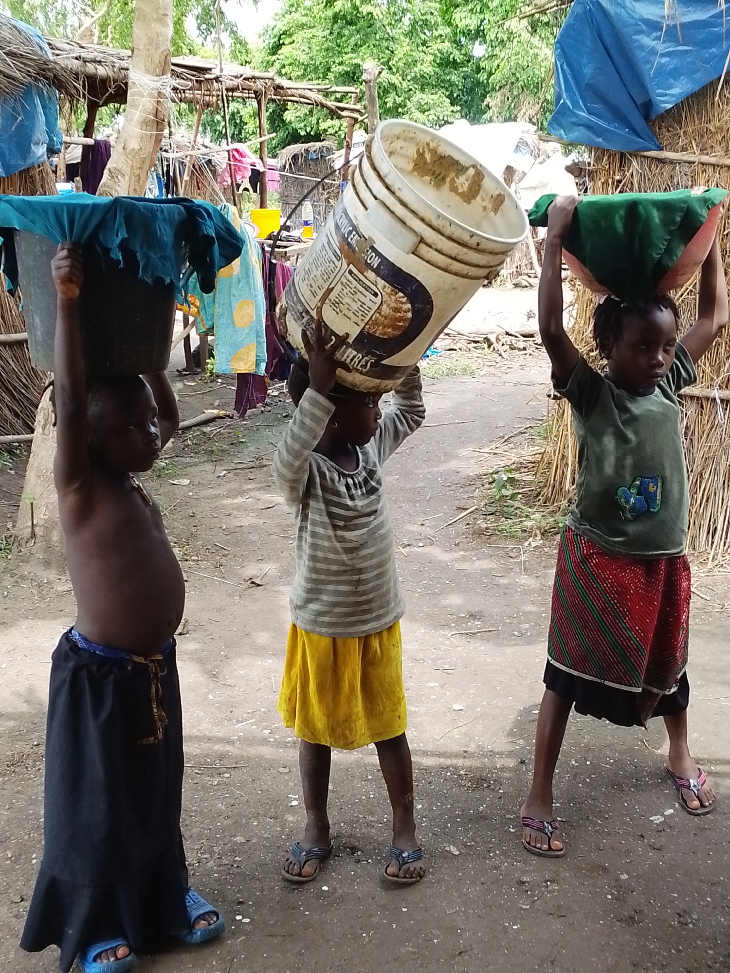 Three young children, two girls and one boy, are carrying heavy loads on their heads in a dirt-floored, open-air environment. The boy, on the left, is shirtless and wearing a long black skirt and blue flip-flops, balancing a large bucket covered with a blue cloth. A girl in the center wears a striped long-sleeved shirt and a yellow skirt, carrying a bucket covered with a green cloth. To the right, another girl, dressed in a green t-shirt and a red and green striped skirt, carries a pink and green bundle on her head. Behind them, rustic structures made of thatch and branches, along with drying laundry and blue tarps, suggest a temporary or impoverished living situation. The scene depicts a common activity of carrying essential items, likely water or goods, in a rural or informal settlement. The location is identified as Otulu, Nigeria.