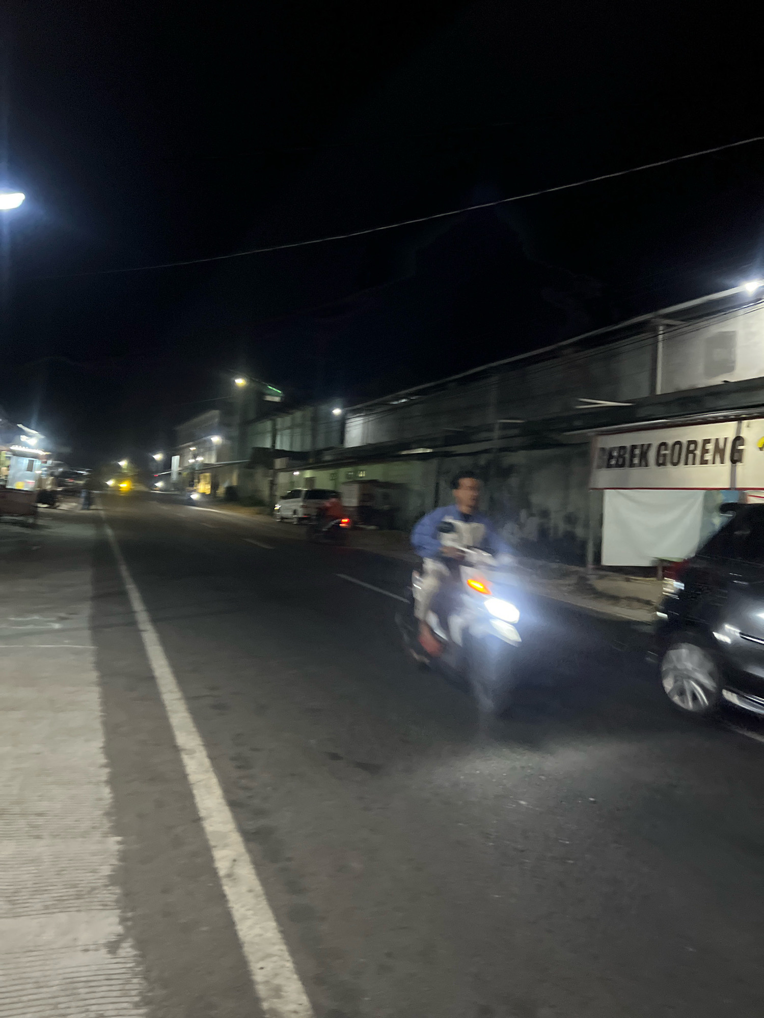 The image depicts a street scene at night in Payaman, Indonesia. A person is riding a motorcycle on the right side of the road, with their headlight illuminated. To the right of the motorcycle, a black car is parked. A banner with the text "BEBEK GORENG" is visible on the side of a building in the background. Other buildings and streetlights are also present, lining the road. The road appears to be paved and has a white line marking its center. The overall scene is characteristic of a commercial street in an Indonesian town at night.