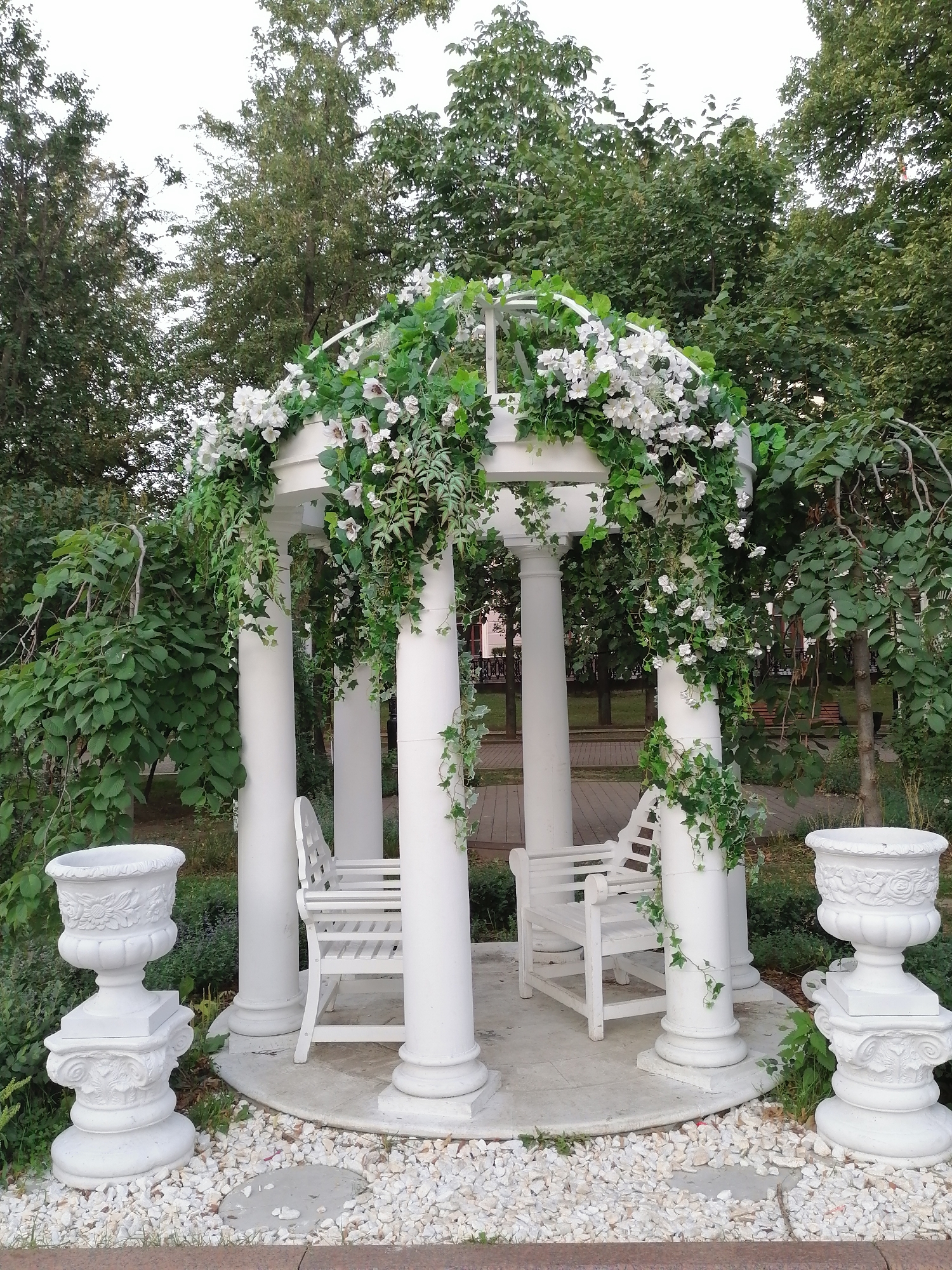 The image shows a white gazebo decorated with artificial white flowers and green vines in a park setting. The gazebo has four white columns and is adorned with an archway also covered in greenery and flowers. Inside the gazebo, there are two white benches. In front of the gazebo, on the left and right sides, are two white decorative urns. The ground around the gazebo is covered in white gravel with some grey stones interspersed. The background consists of lush green trees and foliage, suggesting a park or garden environment. The overall impression is one of a picturesque and well-maintained outdoor space, likely intended for relaxation or events. The image does not contain any people or indicate a specific city, let alone Moscow, Russia.