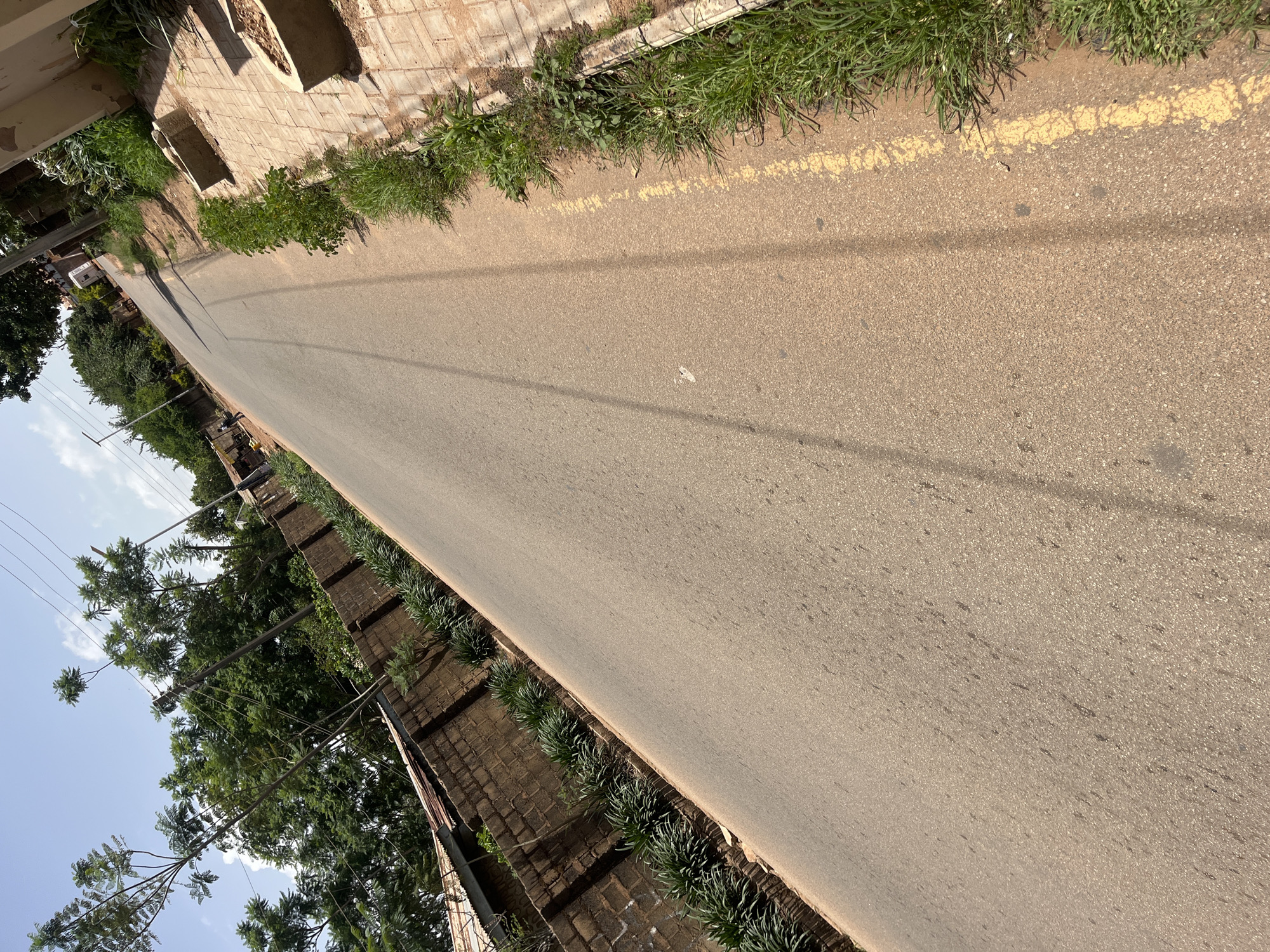 The image depicts a paved road running through a residential area in Gura Top, Nigeria. The road is lined with a brick wall on the left and a sidewalk with grass and some concrete planters on the right. Trees and power lines are visible in the background. The sky is clear and blue. A few scattered dark spots are visible on the road surface.