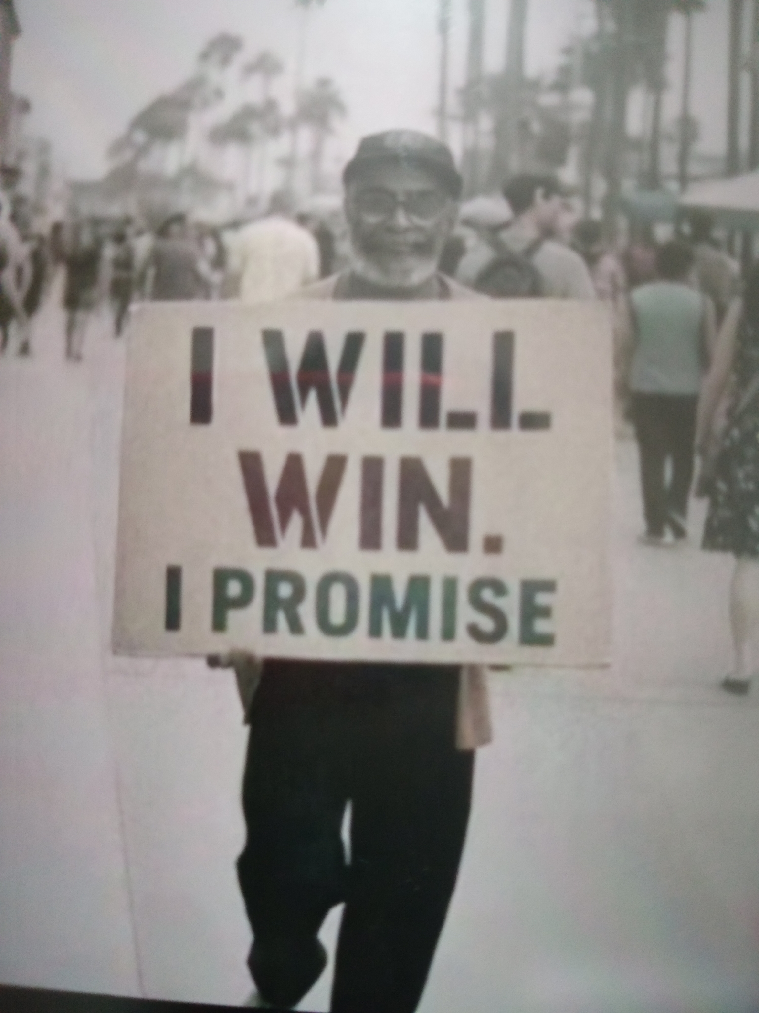 The image shows a middle-aged man holding a sign in a public setting, possibly a street or promenade, with a crowd of people in the background. The man is wearing a hat and glasses and appears to be smiling. The sign he is holding has bold lettering that reads, "I WILL WIN. I PROMISE." The background is blurry, but it appears to be an outdoor environment with palm trees and several individuals walking around. The overall scene is a candid shot, likely captured during the day, given the lighting. There is no visual information in the image to suggest this scene is located in Jalingo, Nigeria.