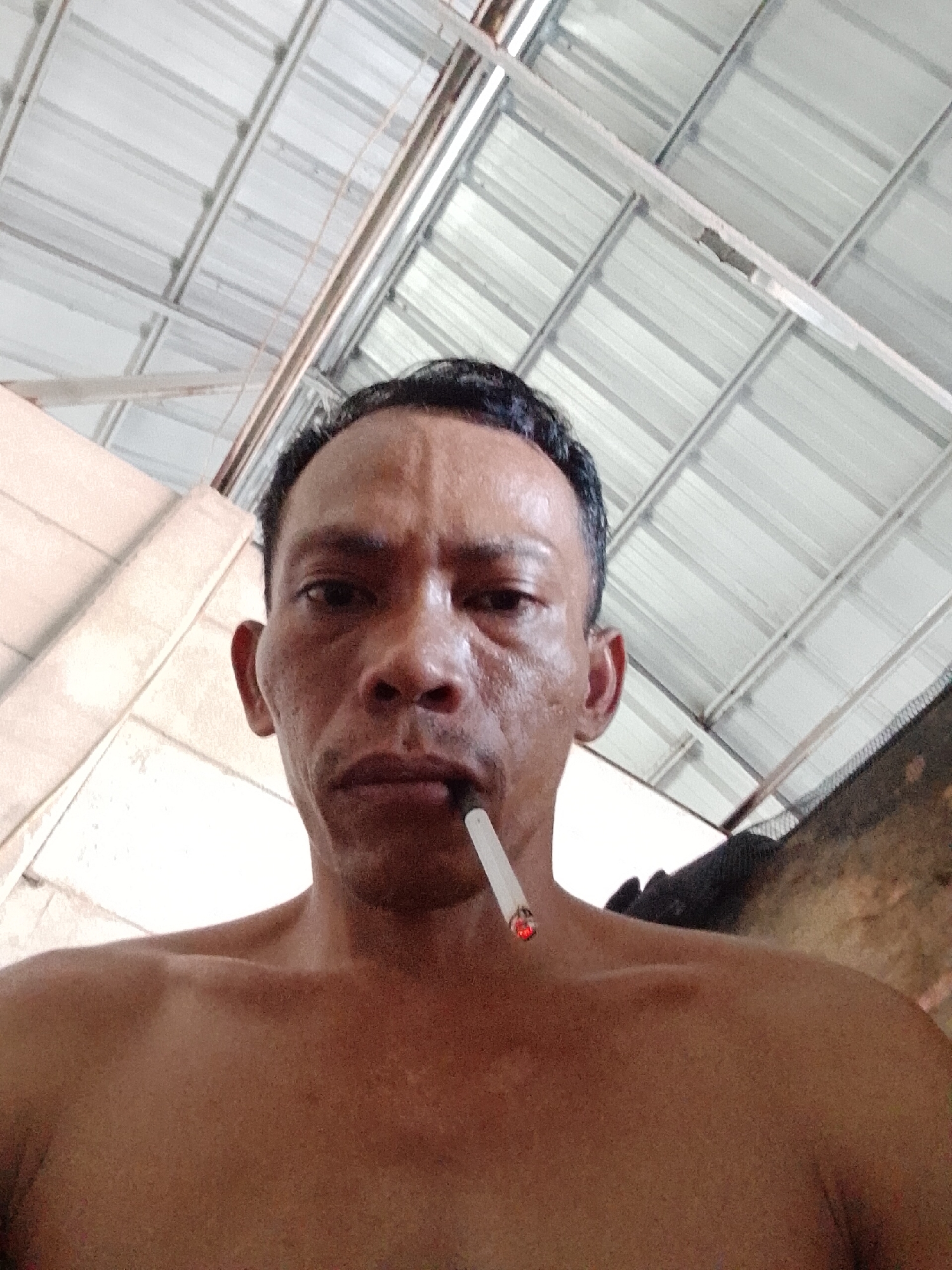 A shirtless, middle-aged Asian man is holding a lit cigarette in his mouth, and is indoors, presumably in Bukit Ketok, Indonesia, with a metal corrugated ceiling above. He is looking directly at the camera.
