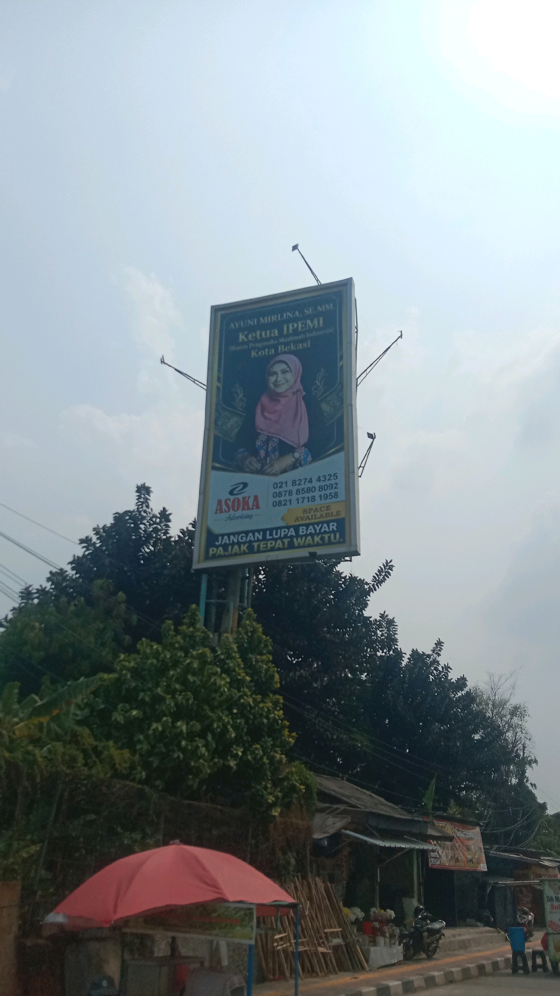 The image shows a billboard in Bekasi, Indonesia, on a cloudy day. The billboard features a portrait of a woman named Ayuni Mirlina, SE, MM, who is identified as the Chairwoman of IPEMI (Ikatan Pengusaha Muslim Indonesia) for the Bekasi region. Below her portrait, there is an advertisement for Asoka Advertising, along with contact numbers and a message in Indonesian that translates to "Don't forget to pay your taxes on time." The billboard is mounted on a tall metal structure and is surrounded by trees and foliage. In the foreground, there is a red umbrella covering a small stall, and a street with a motorcycle parked nearby. The overall scene depicts a typical urban roadside environment in Indonesia.