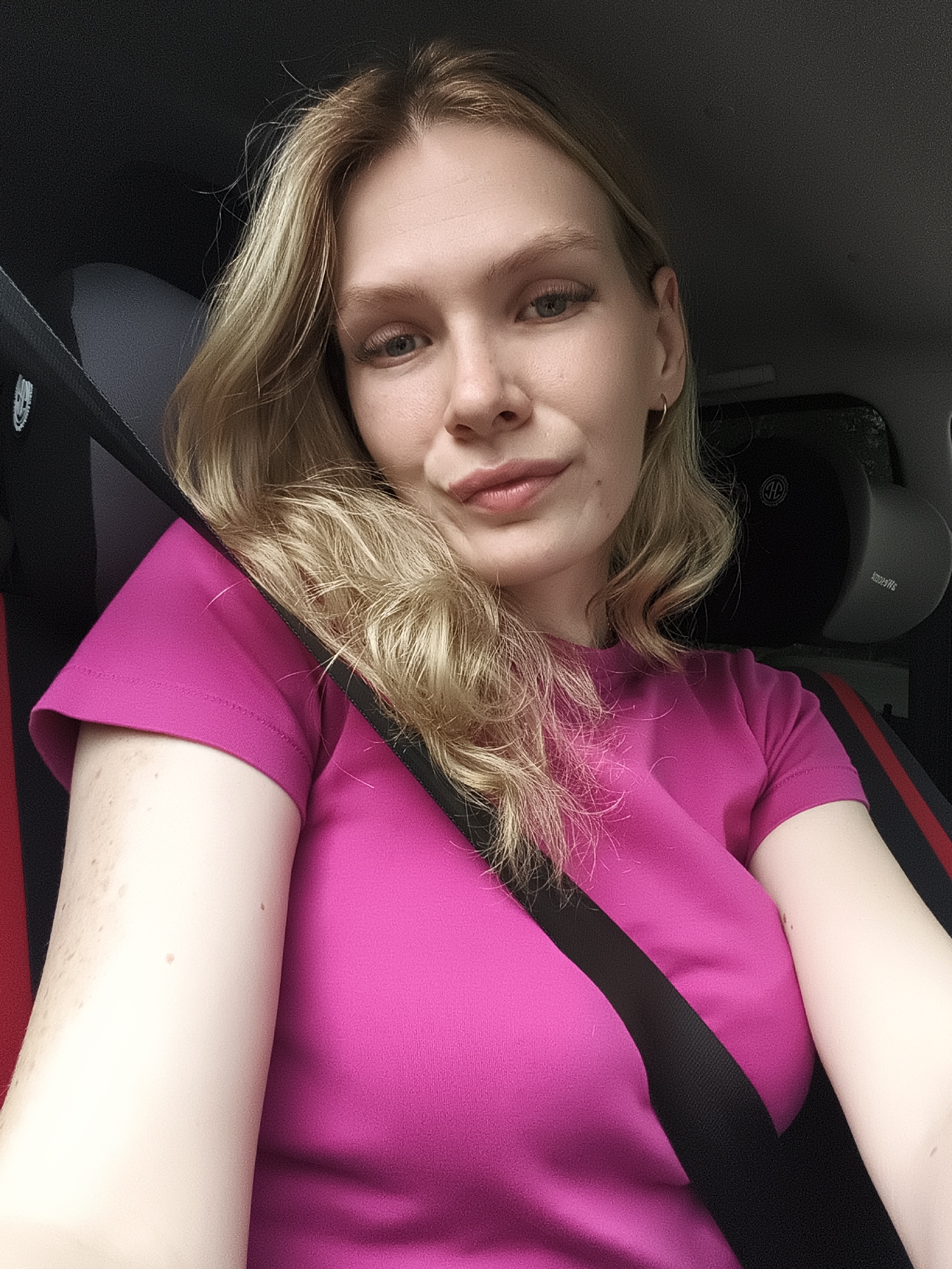 A woman is in the driver's seat of a vehicle. She is wearing a pink shirt and a seatbelt. The interior of the car has black and red accents. A headrest is visible with an emblem. The background is blurred. There is no information in the image to determine a location in Izhevsk, Russia.