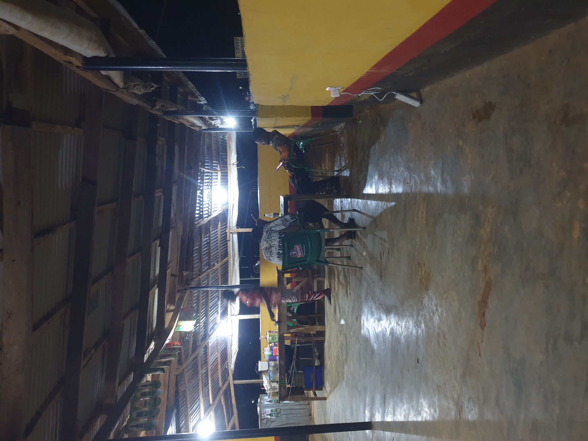 This is a nighttime, medium shot of an open-air establishment in Anilelerin, Nigeria. The structure has a corrugated metal roof supported by wooden beams and dark metal posts. Bright fluorescent lights illuminate the scene, casting strong reflections on the concrete floor, which appears damp and shows some staining.
In the foreground, a person sits on a green plastic chair facing away from the camera. They are wearing a white t-shirt with a graphic print. To their left, another person is seated in a green plastic chair, looking down at a mobile phone they hold in their hands. This individual is wearing a dark long-sleeved shirt and dark pants.
In the middle ground, a wooden counter or bar area is set up with various items, including bottles and containers. A person in a red and white striped outfit is standing behind the counter, seemingly engaged in activity.
The establishment has a yellow-painted wall with a red stripe near the bottom on the right side. An electrical outlet with a white cord plugged into it is visible on this wall. In the background, the environment is dark, suggesting it is nighttime. The overall impression is a casual, possibly local, gathering place.