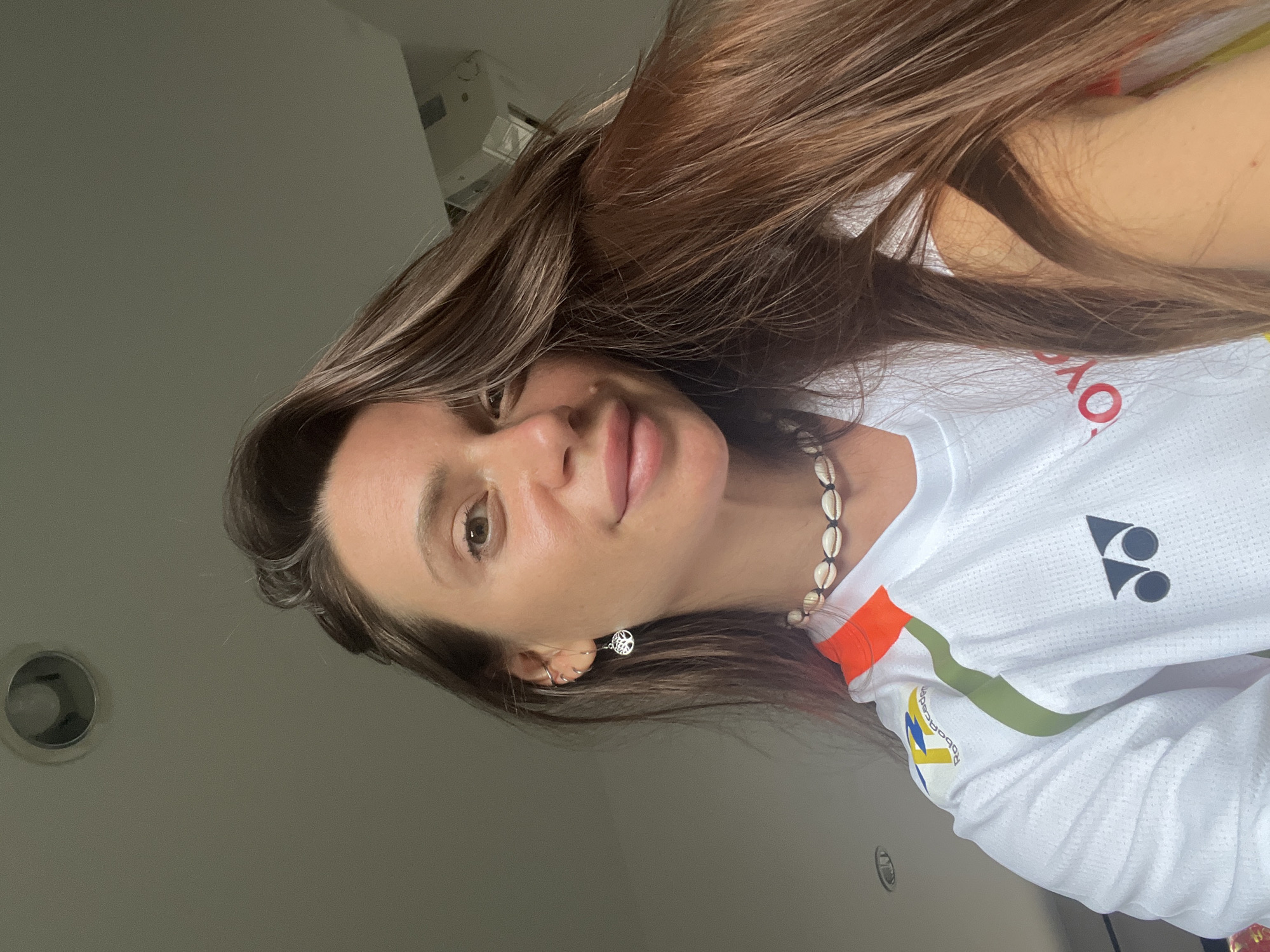 A woman with long brown hair is wearing a white shirt with logos and a shell necklace. She has a peace sign earring in her left ear. The location is Unknown, Unknown.