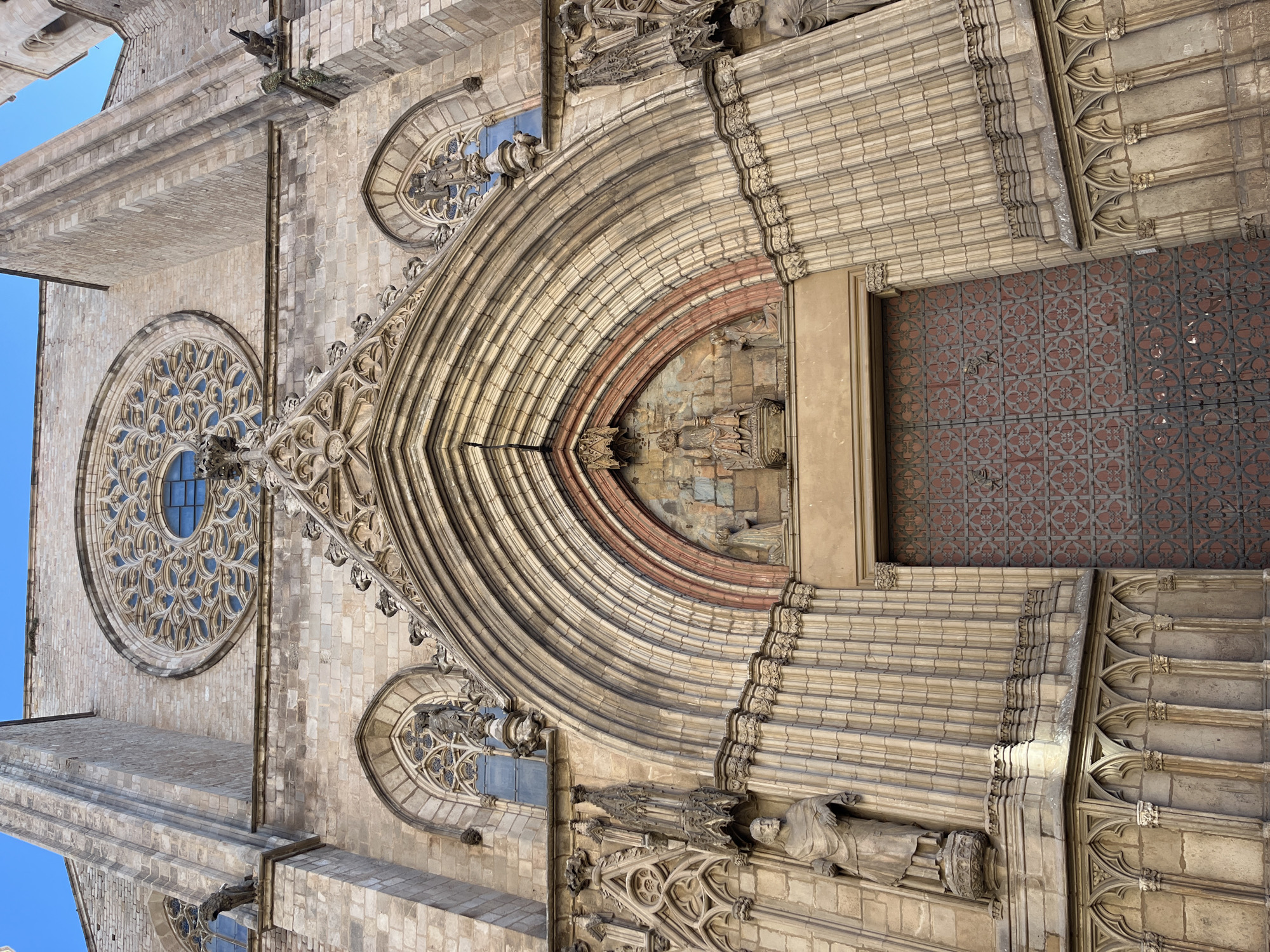 The image shows a detailed view of the facade of the Basilica de Santa Maria del Mar in Barcelona, Spain. The facade features a large rose window on the left, two smaller arched windows, a pointed arch doorway with multiple layers of arches and elaborate carvings. There are stone sculptures flanking the door. The building is made of light-colored stone. The sky, a deep blue, is visible in the upper-left corner of the image. The large door is made of dark brown wood tiles.