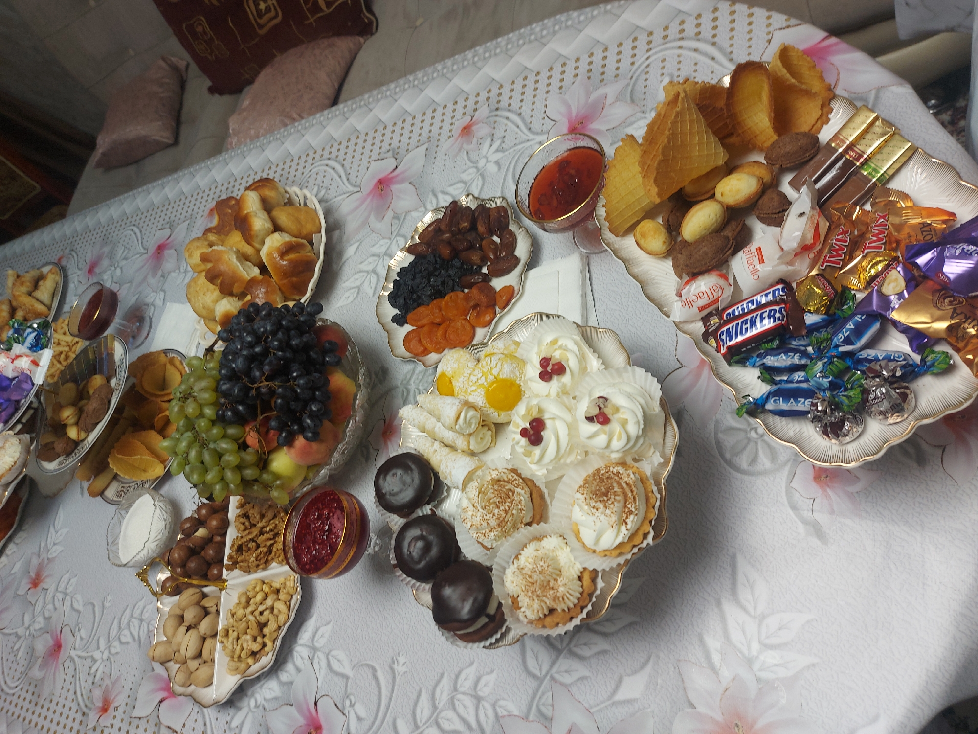 The image depicts a table laden with a variety of snacks and sweets, set on a floral-patterned tablecloth. The spread includes platters of nuts (pistachios, cashews, walnuts, and macadamia nuts), grapes (green and dark purple), dried fruits (apricots, raisins, and dates), pastries, and an assortment of chocolates and candies. There are also small cakes with white frosting and fruit toppings. In the background, a sofa with pillows is visible, suggesting an indoor setting. Based on the provided location of Ақтоғай, Kazakhstan, this scene likely represents a gathering or celebration within a home or a hospitality venue in that region. No people are visible in the image.