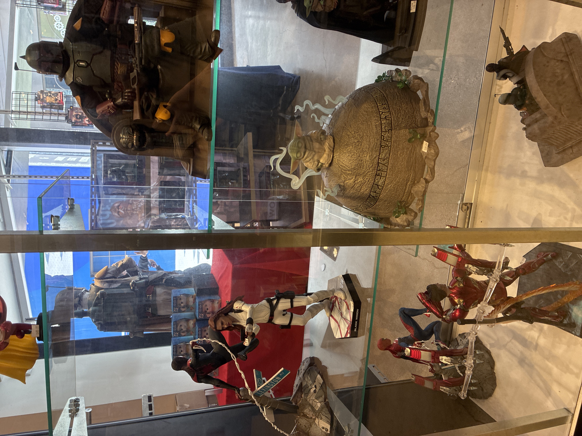 A medium shot shows a glass display case filled with collectible action figures and statues. The figures are arranged on multiple shelves and pedestals. The top shelf features a large figure of Boba Fett seated on a throne. Below that, on the second shelf, a Black Widow action figure is displayed in a dynamic pose, holding a gun, with a Spider-Man figure in the background. The third shelf showcases a Grogu (Baby Yoda) figure perched on a decorative, weathered dome with an inscription. Further down, on the bottom shelf, a scene with Iron Man and Spider-Man figures is visible, and another group of figures, possibly from The Mandalorian, are on a vehicle. The background is blurred, but glimpses of shelves, posters, and lighting suggest an indoor retail environment, possibly a comic book or collectible store. The overall lighting is bright, illuminating the glass case and its contents. The location is identified as Unknown, United States.