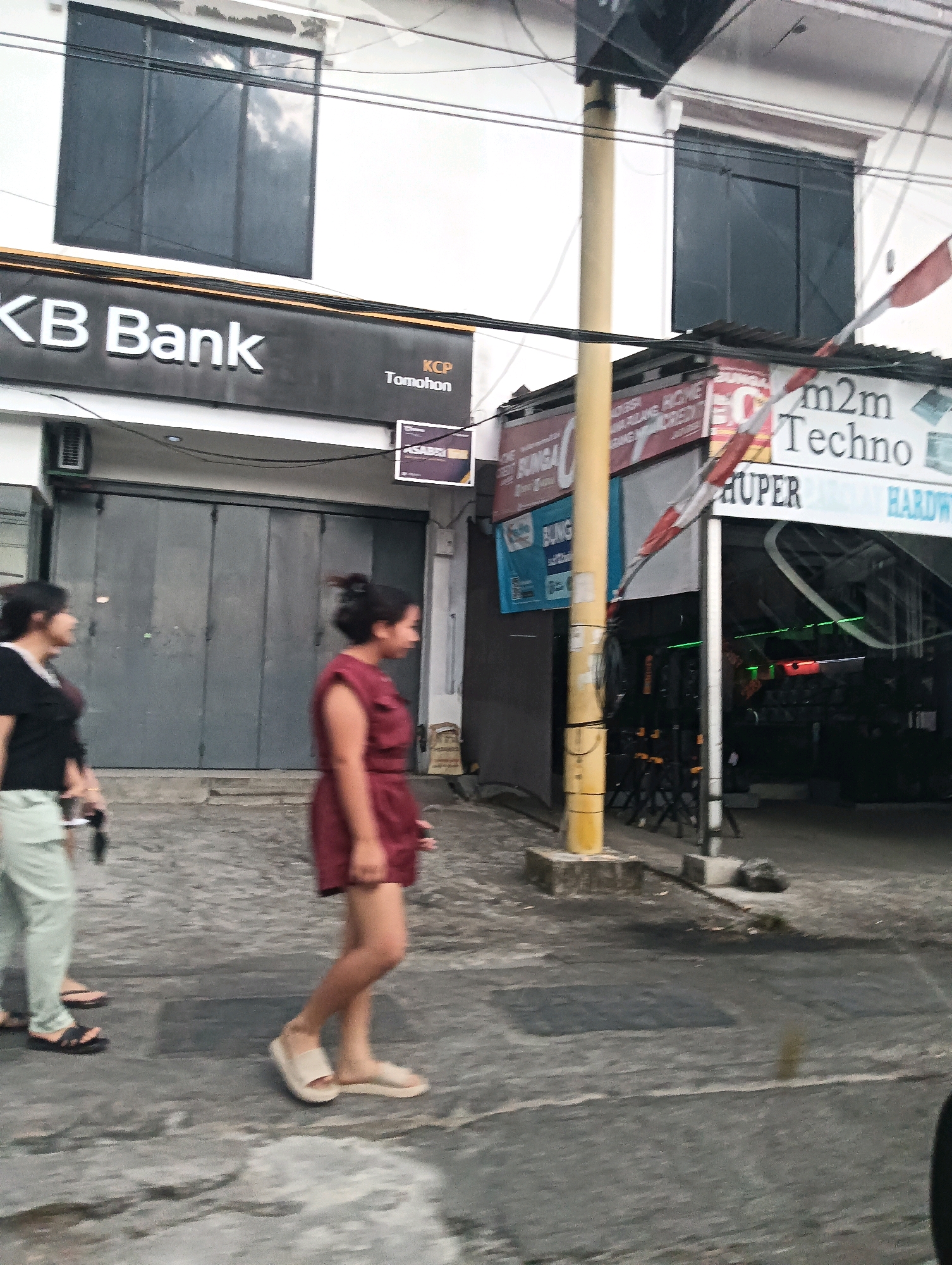 The image captures a street scene in Tomohon, Indonesia, on what appears to be a cloudy day. In the foreground, two women are walking on a paved sidewalk. The woman on the left wears a black top and light-colored pants, while the woman on the right is in a reddish-brown dress. To the left of the sidewalk, a building facade is visible, featuring a sign for "KB Bank" and "KCP Tomohon". Adjacent to the bank, another establishment with signs like "m2m Techno" and "HUPER" is partially visible, showcasing items for sale inside, possibly hardware. A yellow pole stands prominently near the establishments. Power lines crisscross the scene overhead. The overall environment suggests an urban setting with commercial activities.