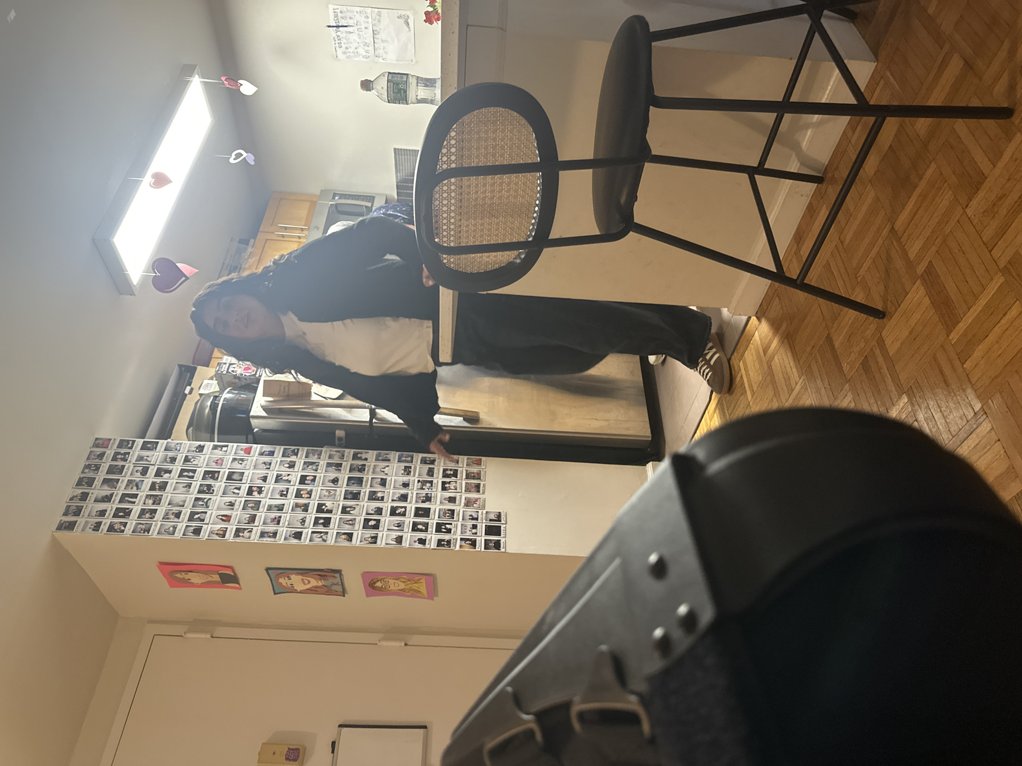 The image appears to be taken inside an apartment or small kitchen. A person wearing a dark-colored sweatshirt and light-colored pants is leaning against a stainless steel refrigerator. A black stool with a woven seat is to the right of the person. The floor is made of parquet wood. On the wall near the refrigerator are numerous small, square photographs. Below them, on the wall near a door frame, are three drawings/sketches of women's faces. The foreground is dominated by a large black object, possibly part of a piece of equipment, obscuring the lower portion of the image.
Without additional information, it's impossible to confirm the location is in New York City. However, the general architectural style of the apartment, and the dense arrangement of photographs on the wall, are suggestive of urban living in a place like NYC.