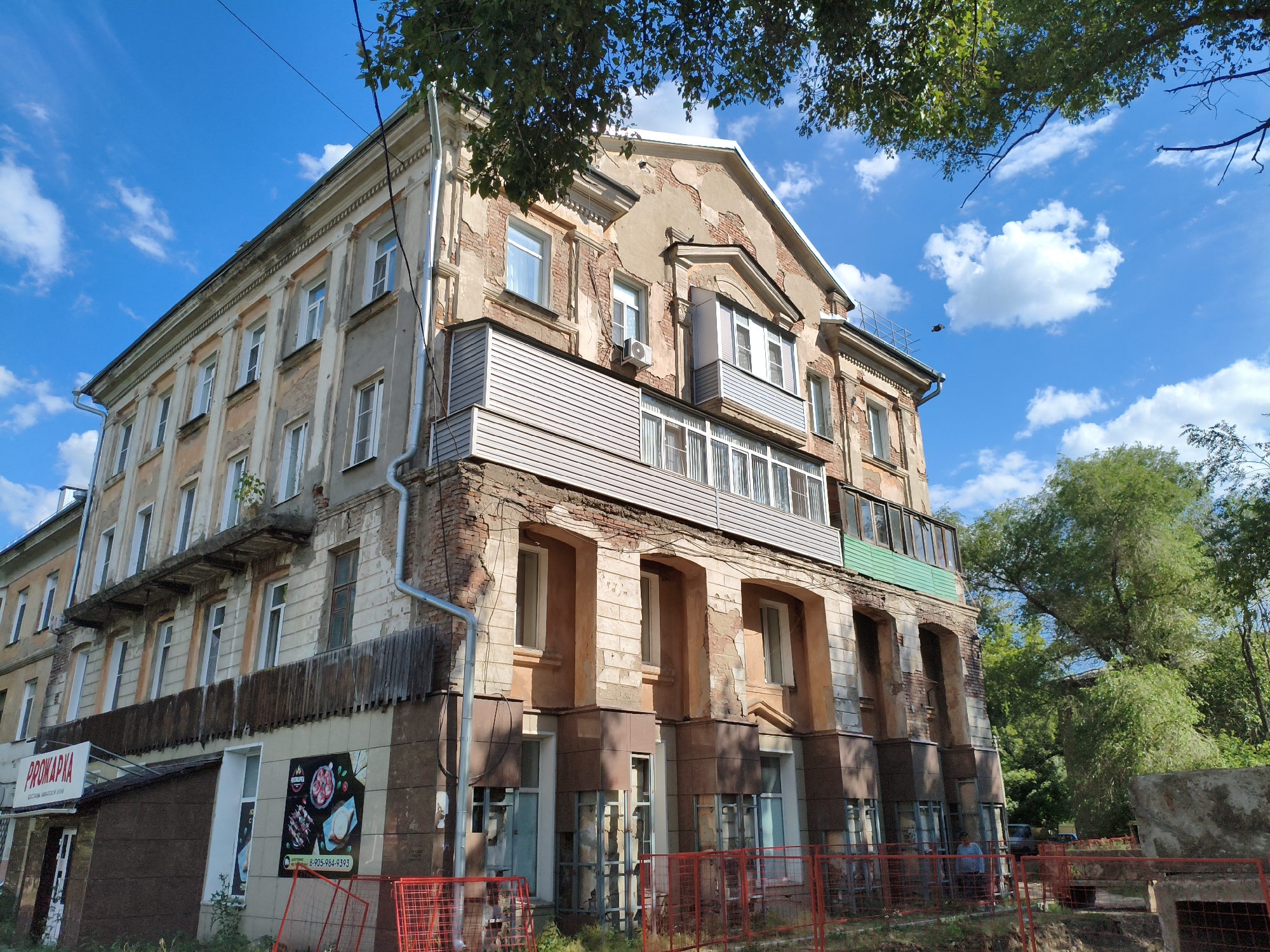 This media file depicts an exterior view of a multi-story building under a bright blue sky with scattered white clouds. The building appears to be aged, with some areas of peeling paint and exposed brickwork, suggesting it might be in need of renovation. The architecture includes multiple levels of windows, some with balconies or enclosed porch areas. There are trees visible in the foreground and background, with foliage casting shadows on the building and surrounding area. Red metal fencing is present in the foreground, indicating potential construction or a protected zone. A sign with "PROMARKА" is visible on the lower left, and another sign with food imagery and a phone number is on the lower right. Two people are visible in the distance, near the right side of the frame, partially obscured by the fence and greenery. The overall scene suggests an outdoor daytime setting, possibly in a residential or mixed-use urban area. The lighting indicates it is daytime. The weather appears to be clear and sunny.