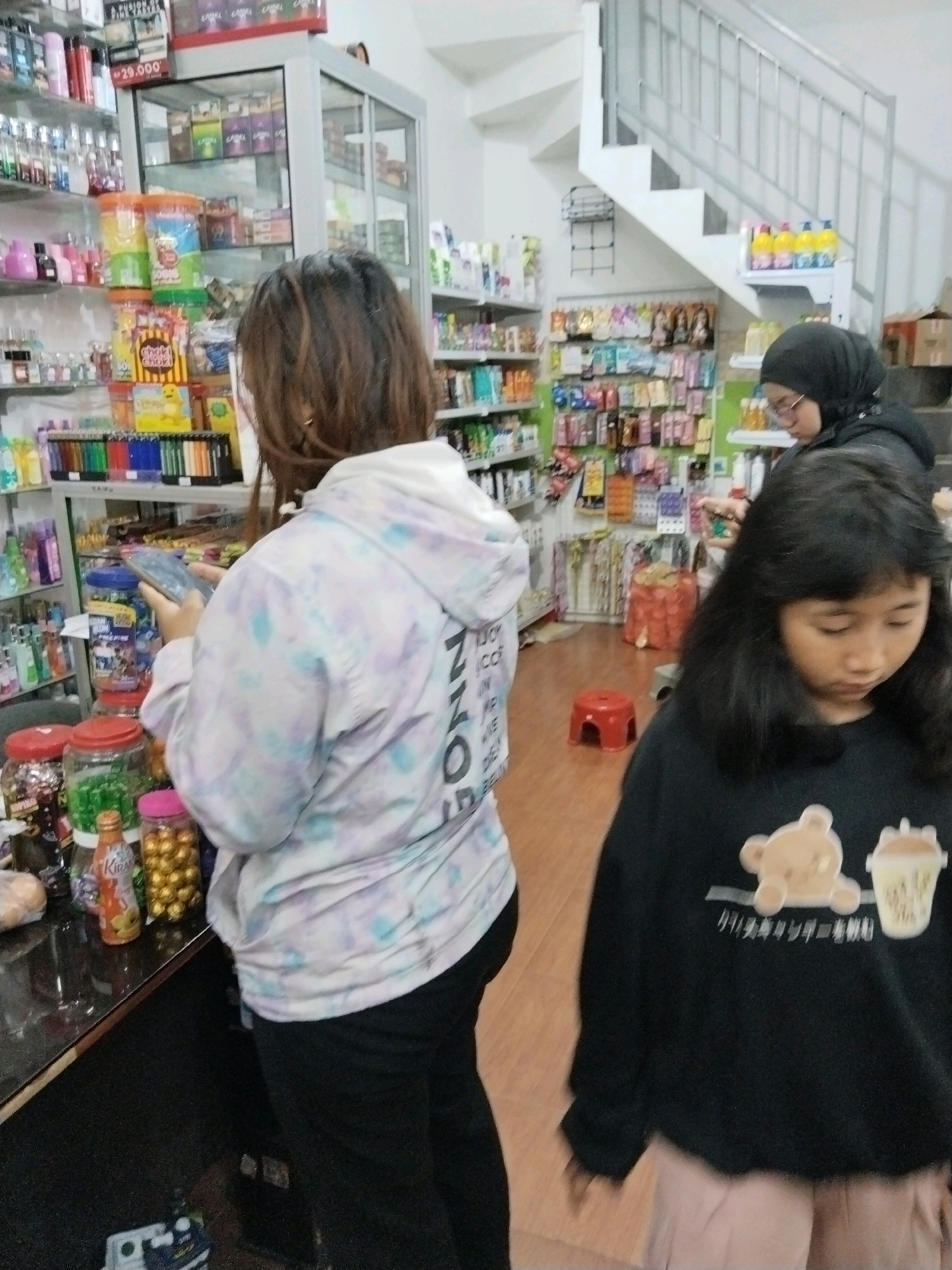 In the city of Banjararum, Indonesia, an indoor convenience store is depicted. The scene features three individuals: two young girls and a woman. One girl, wearing a black long-sleeved shirt with a teddy bear and cup graphic, is positioned in the foreground, looking downwards. Behind her, a second girl is partially visible, wearing a pastel tie-dye hooded jacket and black pants, and looking at her phone. A woman in a black hijab and glasses stands further back, her gaze directed towards the shelves. The store's interior showcases numerous shelves stocked with various products, including snacks, beverages, and personal care items. A staircase is visible in the background, leading to an upper level. The lighting is artificial, and the overall atmosphere suggests a typical retail environment.