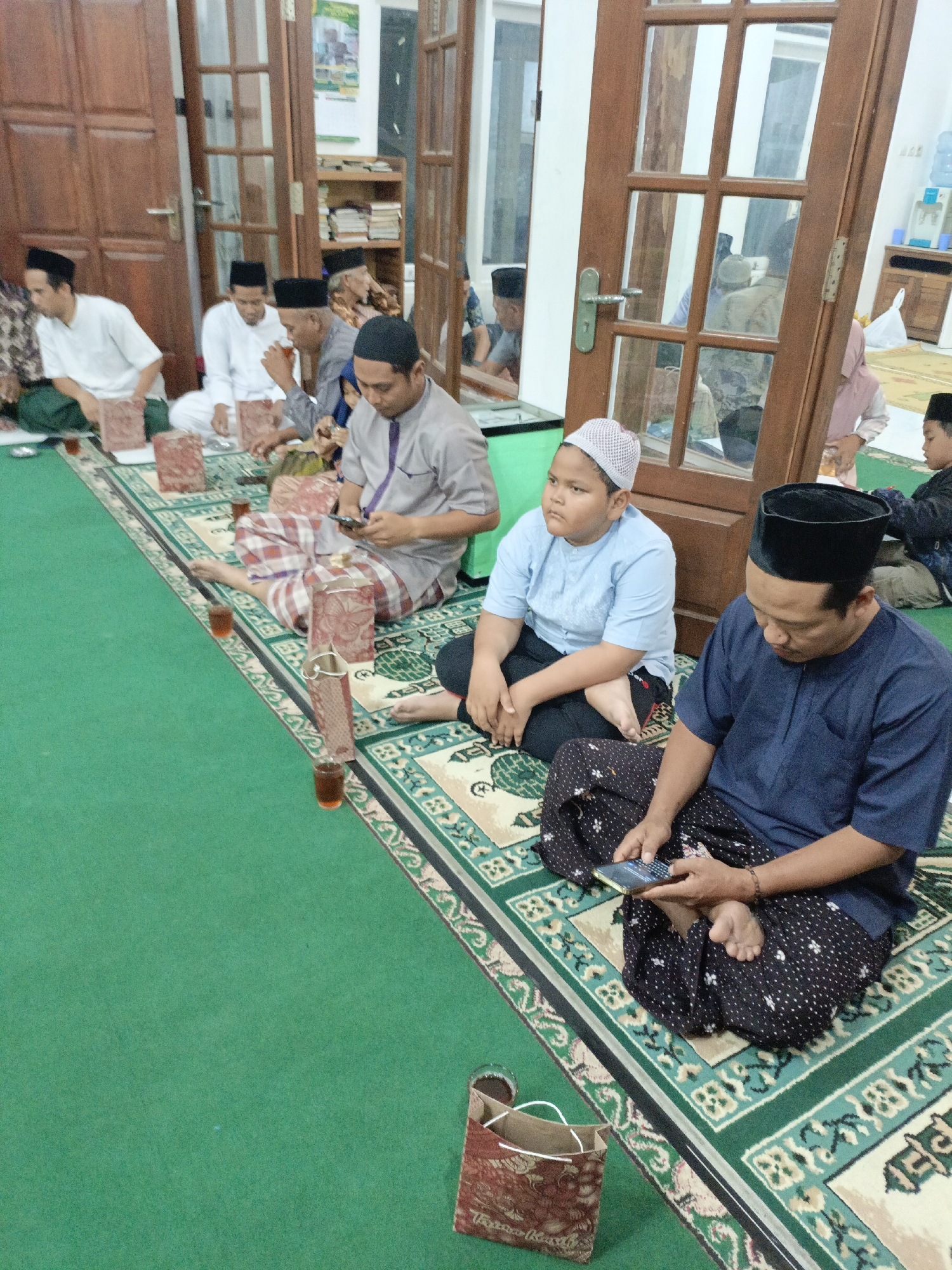The image depicts an indoor gathering of approximately ten people, predominantly men and boys, seated on a green carpet with an Islamic patterned rug. They are dressed in traditional attire, including peci (caps) and sarongs. Several individuals are holding or looking at mobile phones, suggesting a moment of quiet activity or engagement with technology. In the background, wooden doors and shelves stocked with books are visible, indicating a room that could be a mosque, a community hall, or a home. The presence of small glasses of amber liquid, possibly tea, suggests refreshments are being served. The overall atmosphere appears calm and communal. Based on the visual cues and common architectural styles, this scene is likely located in Salatiga, Indonesia.
