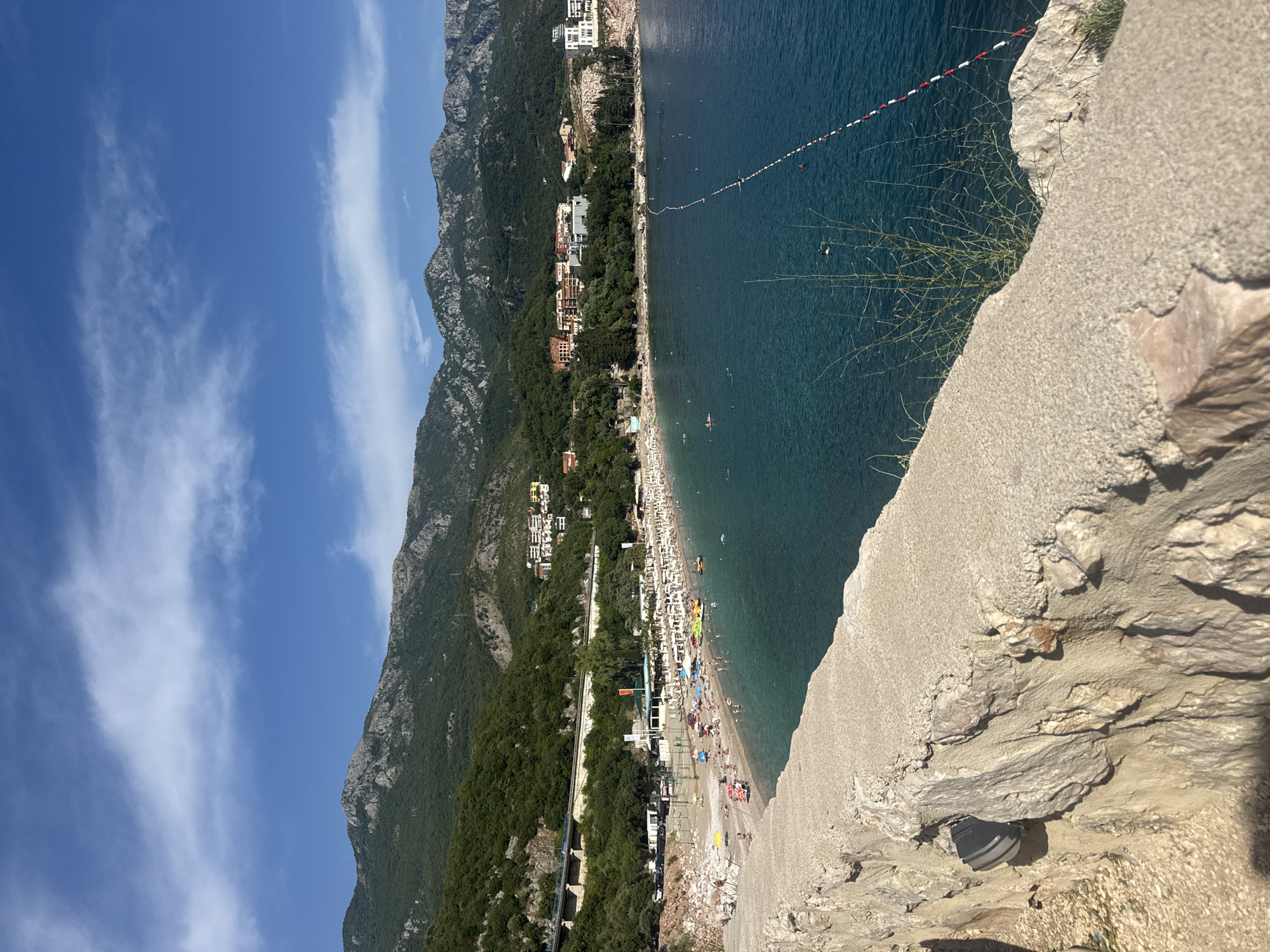 This is a landscape photograph taken from a high vantage point overlooking a crowded beach on a sunny day. The image is oriented vertically.
The scene depicts a wide, pebble beach lined with rows of white sun loungers and umbrellas. Numerous people are visible on the beach, some relaxing, others walking or swimming in the clear, blue sea. A red and white marker buoy line extends across the water, indicating a designated swimming area.
Behind the beach, a densely forested mountainside rises steeply, dotted with buildings, suggesting a coastal town or resort. The architecture appears modern, with several multi-story white buildings visible amongst the trees. A road runs parallel to the beach, separating it from the forest.
In the foreground, a rough, rocky ledge occupies the bottom right of the frame, with a pair of dark-colored shoes visible among the rocks. The sky above is a vibrant blue, with thin, wispy white clouds scattered across it. The overall impression is one of a bustling, sunny vacation destination. The time of day appears to be midday or early afternoon, given the bright sunlight and shadows. The location is identified as Kamenovo, Montenegro. No visible text is present in the image.