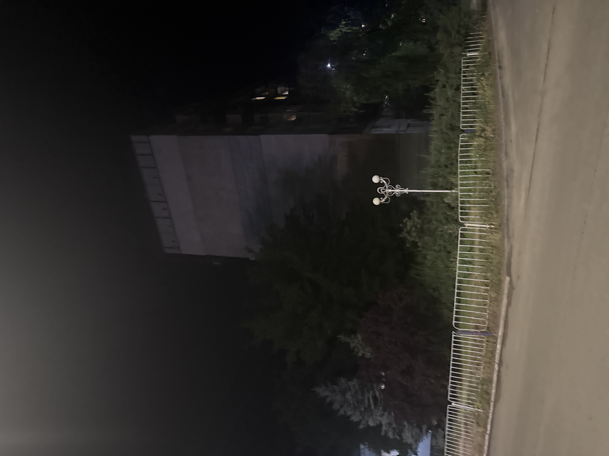 The image shows a building at night, with trees and a fence in the foreground. The building is tall and appears to be an apartment building, with lights on in some of the windows. The fence is white and made of metal posts. There is a lamppost in the middle of the image. The scene is dark, with the building and trees silhouetted against the night sky. The location is identified as Dnipro, Ukraine.