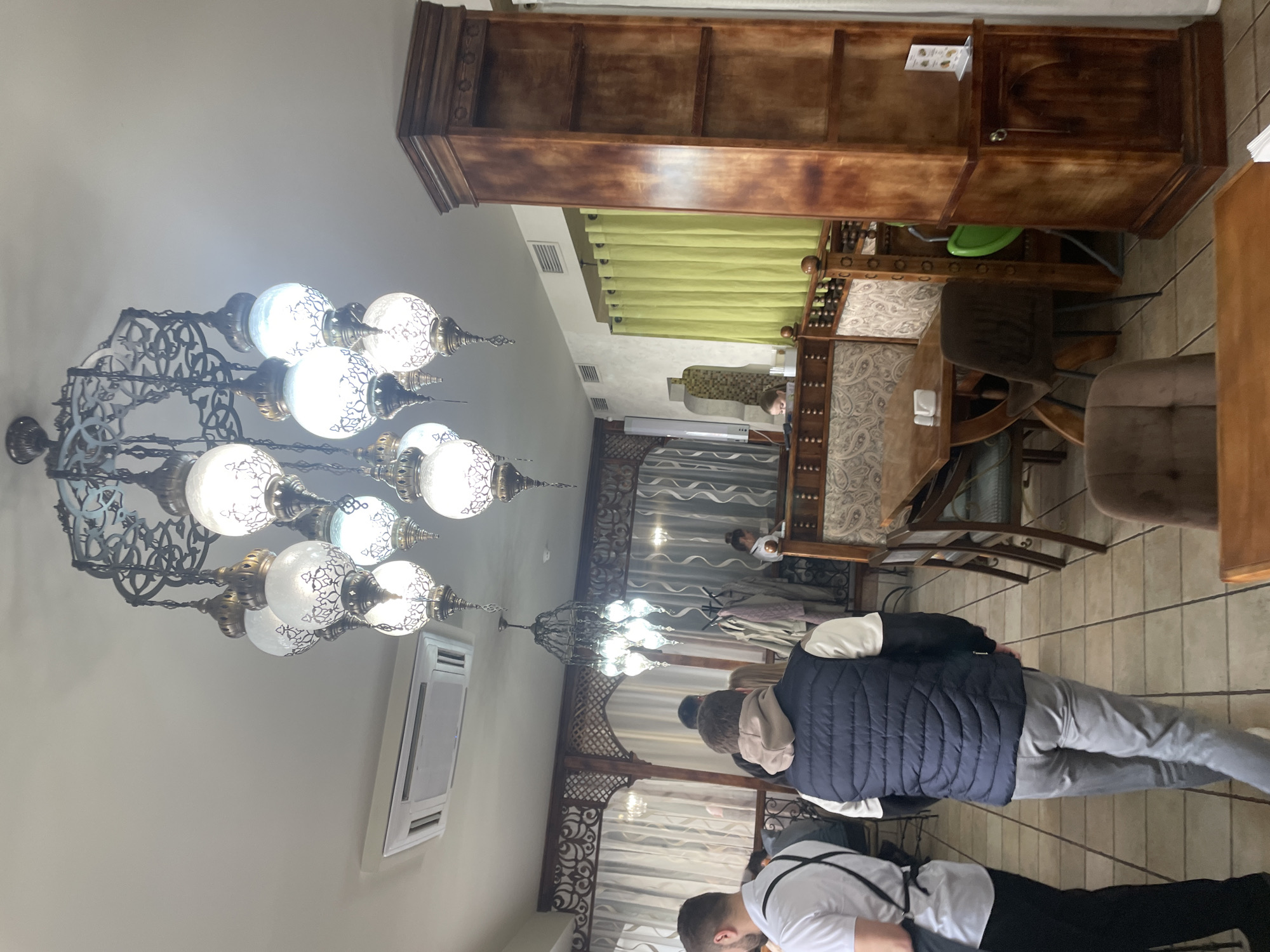 A view inside an establishment in Perm, Russia shows a large, decorative chandelier hanging from the ceiling. The lighting fixture features multiple glass orbs with ornamental designs. Part of an air conditioning unit is mounted on the wall. A wooden cabinet or shelving unit sits atop patterned chairs and tables in the background. Patrons are present in the room, including a person in a navy vest and gray pants, and another person wearing a white shirt and black pants. There is a decorative wooden frame in the back, also with ornamental designs.