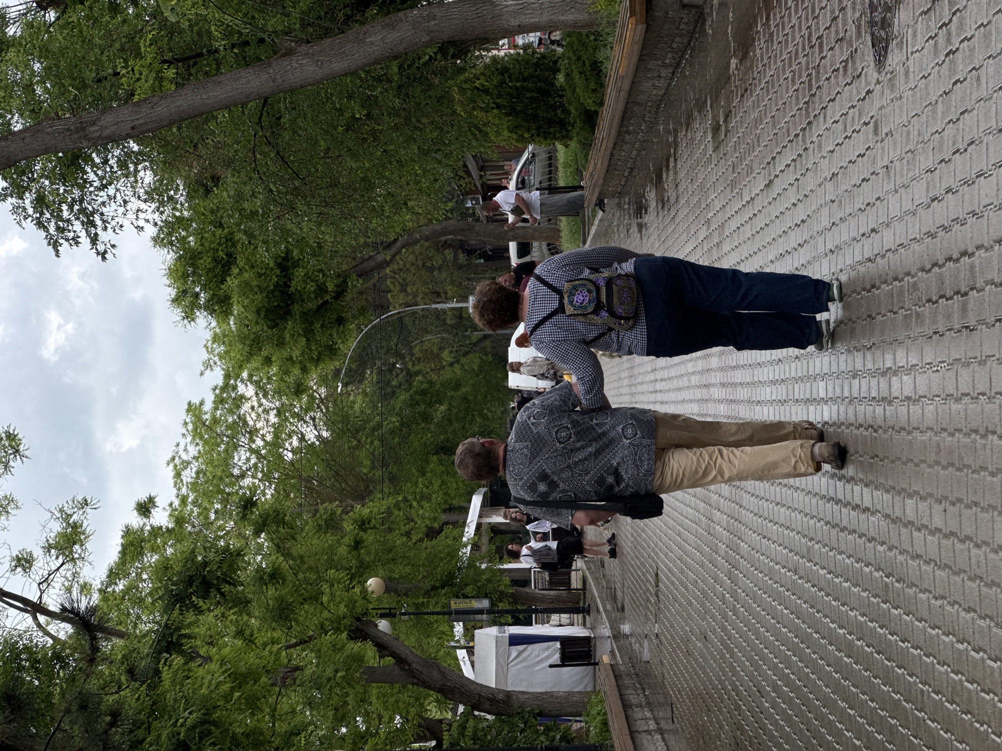 Two people are walking on a brick sidewalk in Yevpatoriya, Ukraine. One person is wearing a blue and white checkered shirt and blue pants, and has a patterned bag on their back. The other person is wearing a gray paisley shirt and tan pants. They are walking away from the camera. In the background, there are trees, street lamps, and white tents. There are other people walking around in the background. The sky is overcast.