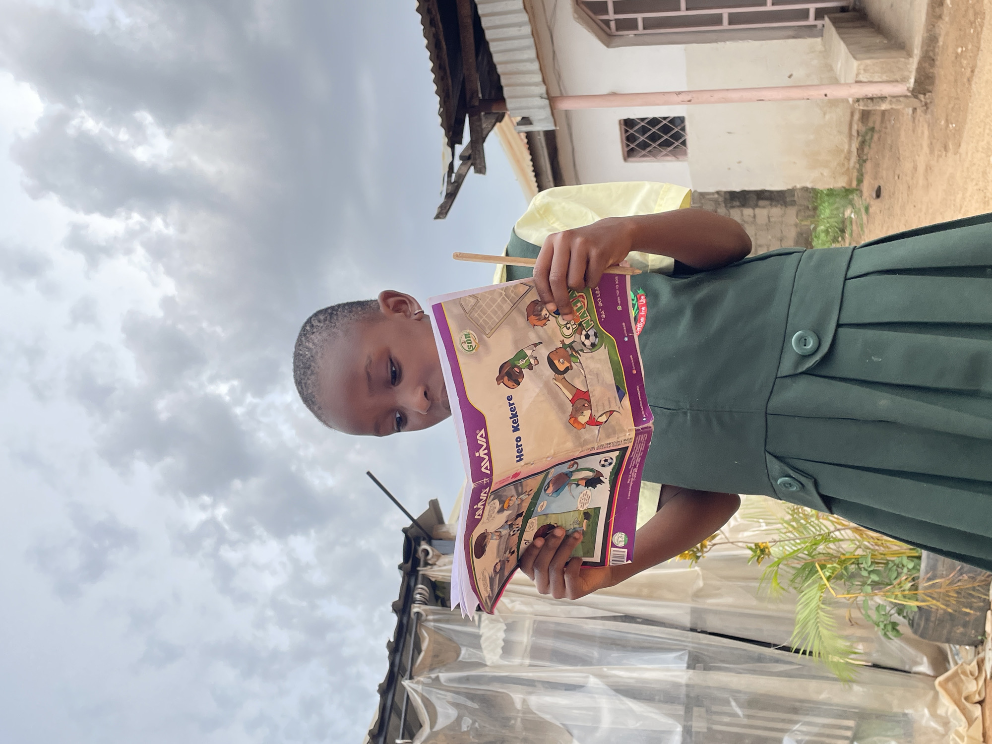 A young girl in a yellow-collared, dark green school uniform stands outdoors reading a purple and white comic book titled "AVIVA Hero Kekere." The book features cartoon illustrations of children playing soccer and other activities. The girl holds a wooden stick in her left hand, potentially used for pointing or keeping her place in the book.
The background includes a building with a corrugated metal roof, a white wall with a small barred window, and a cloudy sky. There are plants and what appears to be a greenhouse or covered structure partially visible.
Given the presence of "AVIVA" branding on the book and contextual clues, the location can be inferred as Jos, Nigeria, where AVIVA Foundation is known to be involved in educational initiatives.