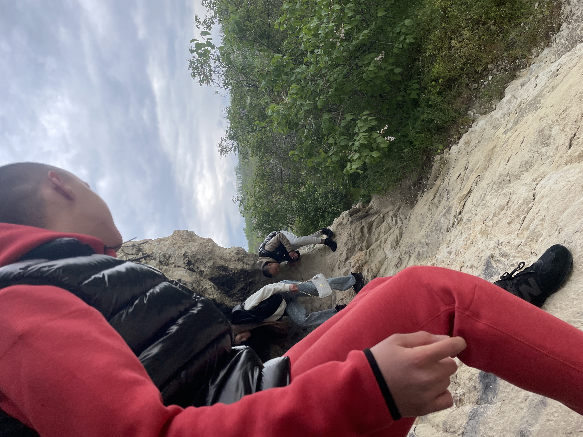 This outdoor image captures a low-angle perspective of three individuals on a rocky, vegetated hillside under an overcast sky.
In the foreground, a person is partially visible, facing away from the viewer but looking upwards and to the left. They wear a red hooded sweatshirt and a black puffy vest. Their head, with a shaved hairstyle, is seen in profile. Their right arm, clad in red, is bent, with a hand visible, wearing a black wristband. On the bottom right, a black athletic shoe with a white sole and a prominent "N" logo, likely from New Balance, rests on the light-colored, rough rock.
Further up the incline, two other people are present. One individual, positioned slightly to the left, wears a white long-sleeved top, a black vest, and light blue jeans. They are holding a white quilted purse with a chain strap. Their long, dark hair covers their face. Behind and to the right of this person, another individual in light-colored pants and a dark vest appears to be navigating or resting on the rocky terrain.
The background features dense green foliage, including bushes and trees with small white blossoms, along the left side and top of the image. The sky is overcast with diffused, grey clouds, indicating daytime. The scene suggests a hiking or climbing activity in a natural, rugged environment.