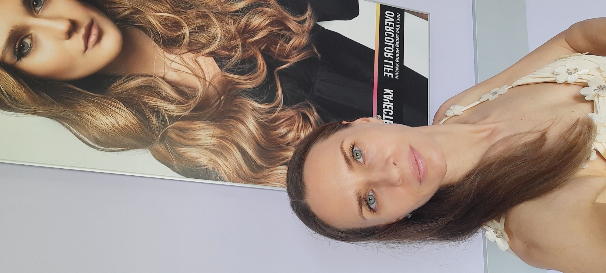 A woman with brown hair and blue eyes is posing in front of a poster featuring a woman with long blonde hair. The poster has text written in Russian, which appears to be a slogan or product name. The woman in the foreground is wearing a light-colored dress adorned with floral embellishments. The background is a plain lavender wall. The image appears to have been taken indoors, possibly in a beauty salon or a commercial space in Moscow, Russia.