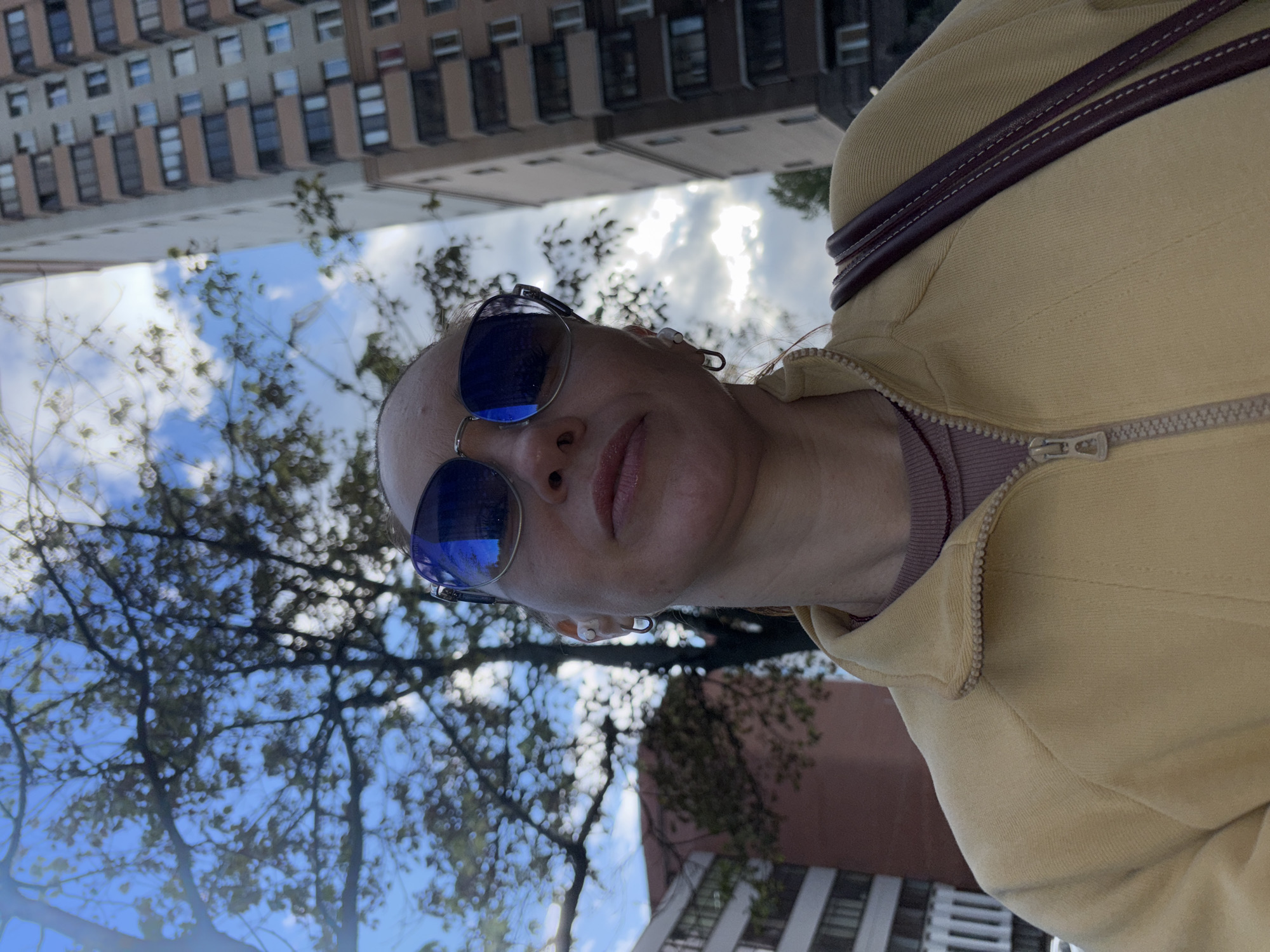 In a low-angle, outdoor, eye-level shot, a woman is pictured from the chest up. She is looking towards the camera, her face partially obscured by large, blue-tinted sunglasses. She has on a light yellow zip-up jacket over a mauve shirt. A dark red purse strap is visible over her left shoulder. In the background, a tall apartment building rises up on the right, and a large tree with sparse green leaves is on the left. The sky is blue with scattered white clouds, suggesting daytime. The scene appears to be in a city.