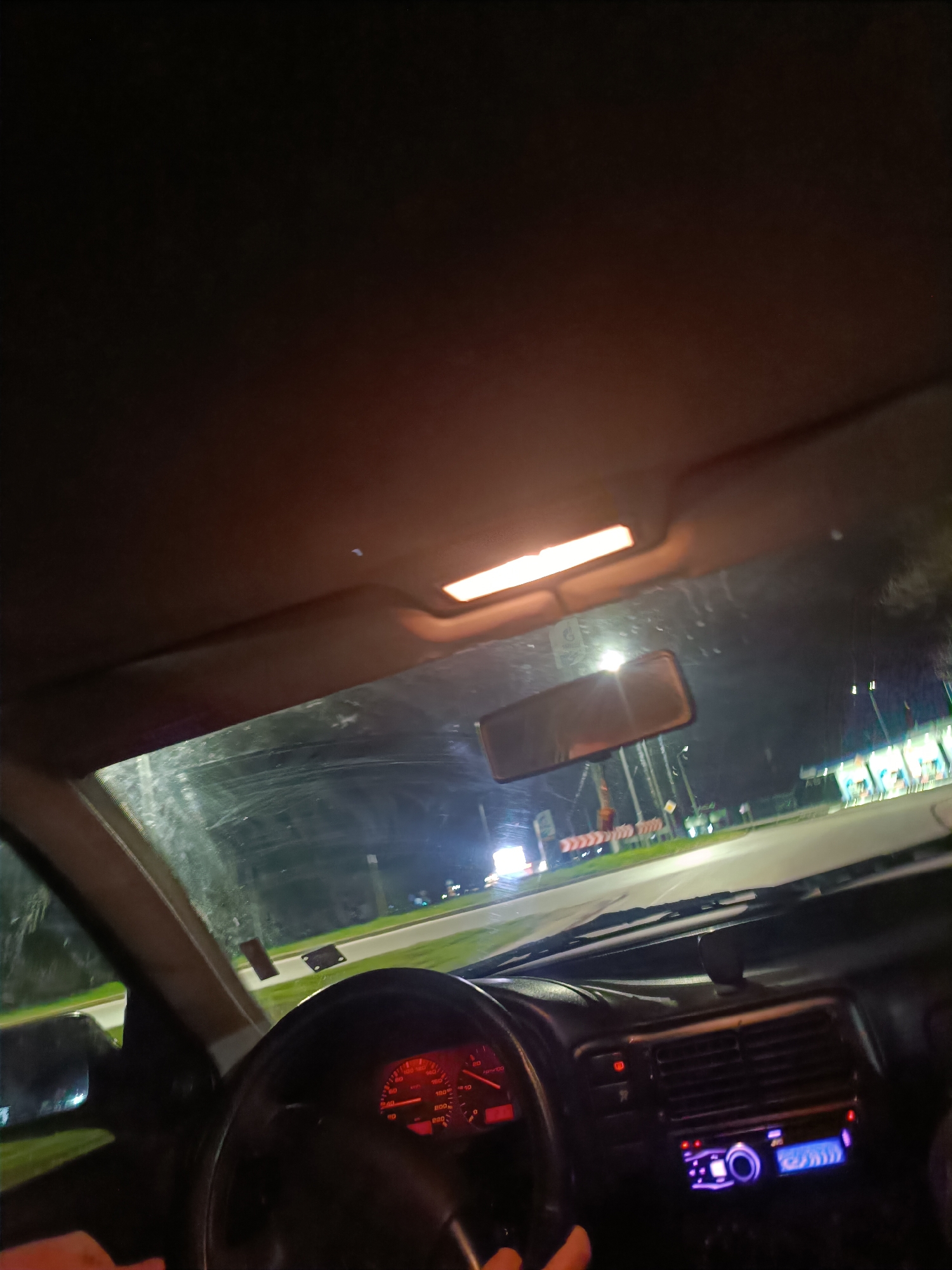 The image shows the interior of a car at night, likely in Plovdiv, Bulgaria. The dashboard is visible, including the steering wheel, speedometer, and a car stereo. The windshield is slightly smeared, with streetlights and buildings visible through it. The rearview mirror is positioned in the center of the windshield. The interior light is on. There's a hand partially visible on the steering wheel. The environment suggests an urban or suburban setting at night.