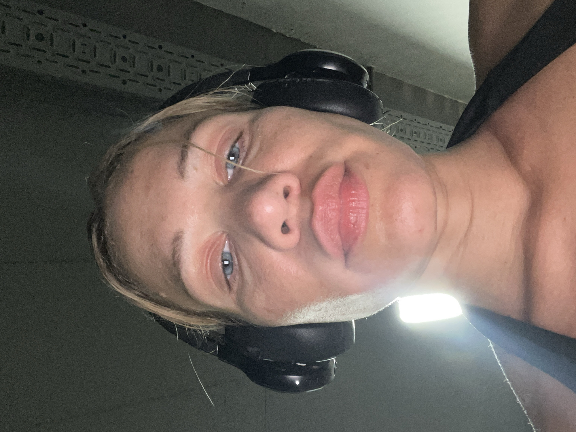 This is a close-up, low-angle selfie of a blonde woman wearing black over-ear headphones and a black tank top. She has blue eyes and her lips are slightly pursed. The background appears to be indoors, with a dimly lit ceiling structure visible, possibly industrial or a gym setting. The lighting is harsh, with a bright light source to her left, casting a strong highlight on her skin. There are no visible landmarks or features that would indicate a specific location within Altötting, Germany.