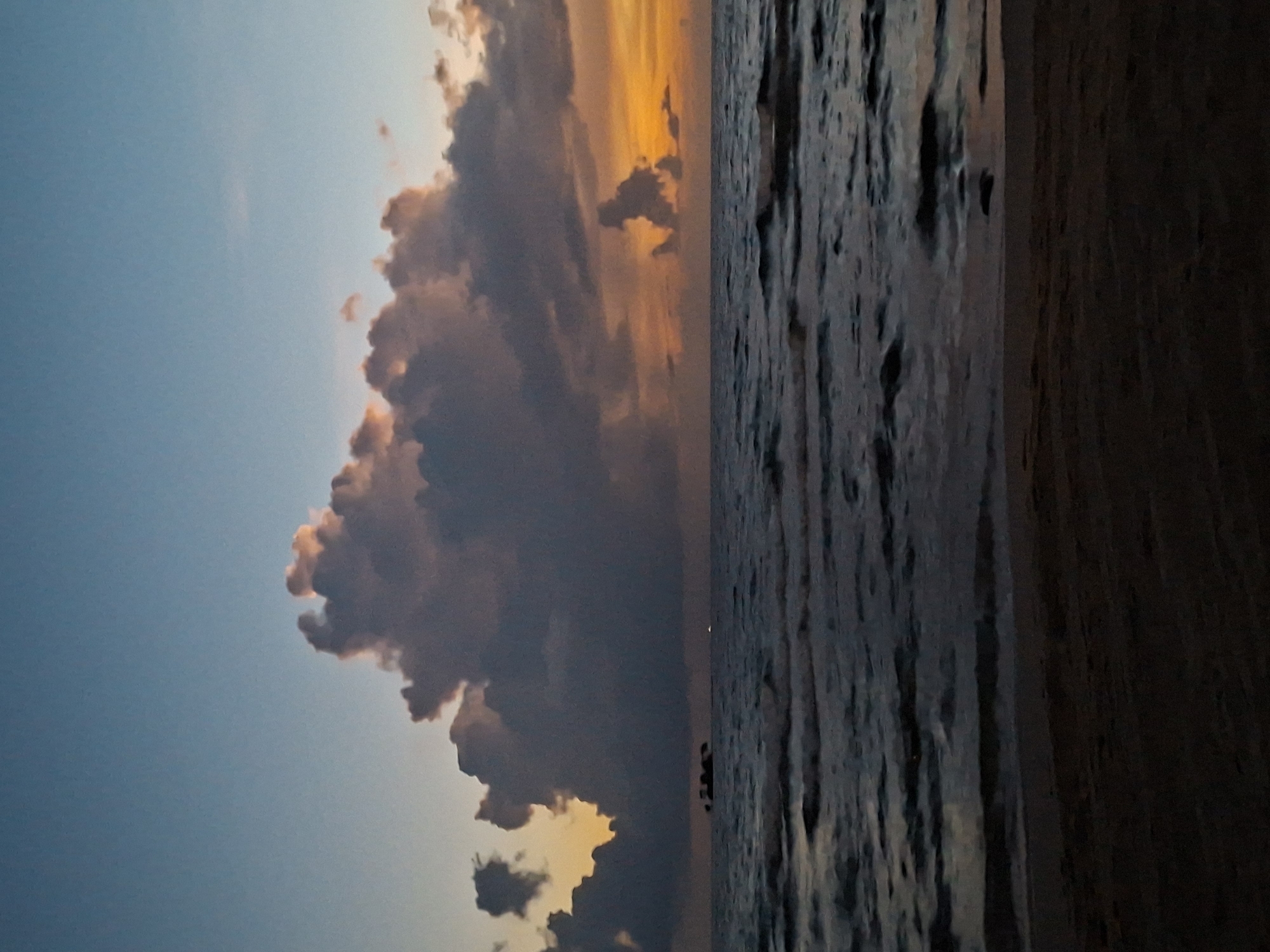 The image shows a beach scene at dusk. The sky is a gradient of blue and orange, with large, dark clouds dominating the upper portion of the frame. The sea is dark and choppy, with small waves lapping at the shore. In the distance, there appears to be a small island or structure in the water. The foreground is a sandy beach, dark and indistinct. The overall mood is tranquil and serene, with the colors of the sunset creating a dramatic backdrop.